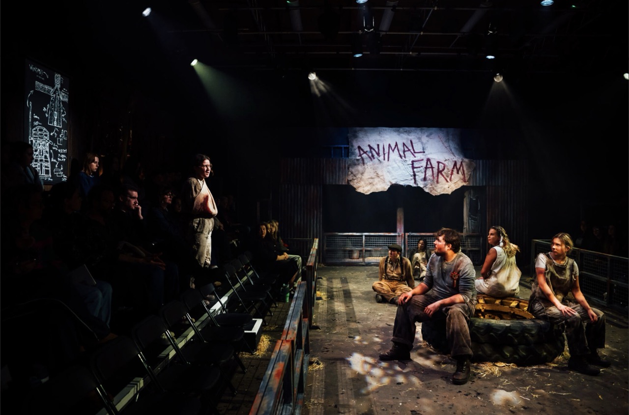 Actors sit on a stage set for Animal Farm, with a painted banner hanging at the back and audience seating along the left side.