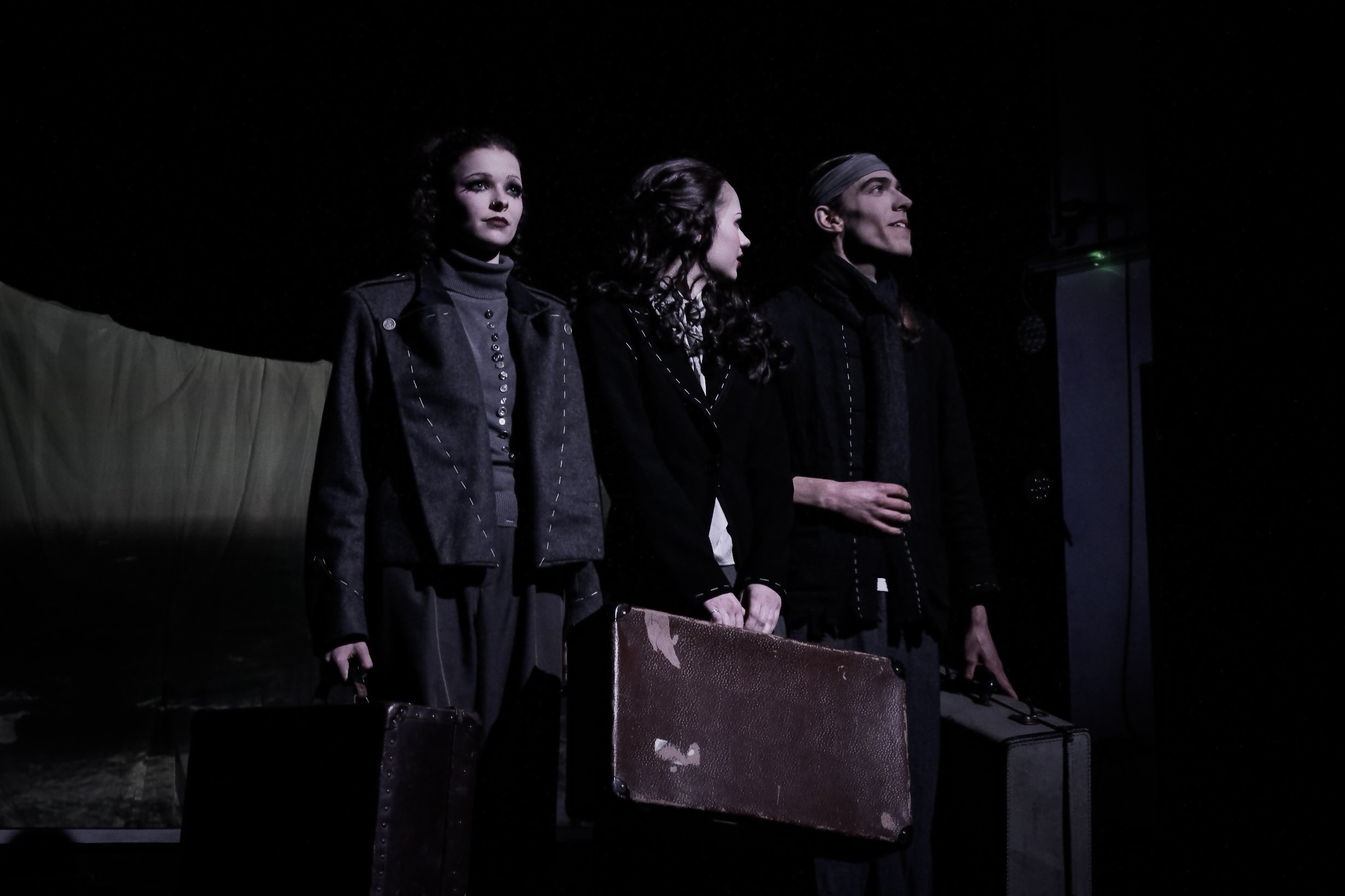 Three adults in dark clothing stand on a dimly lit stage, each holding a suitcase.