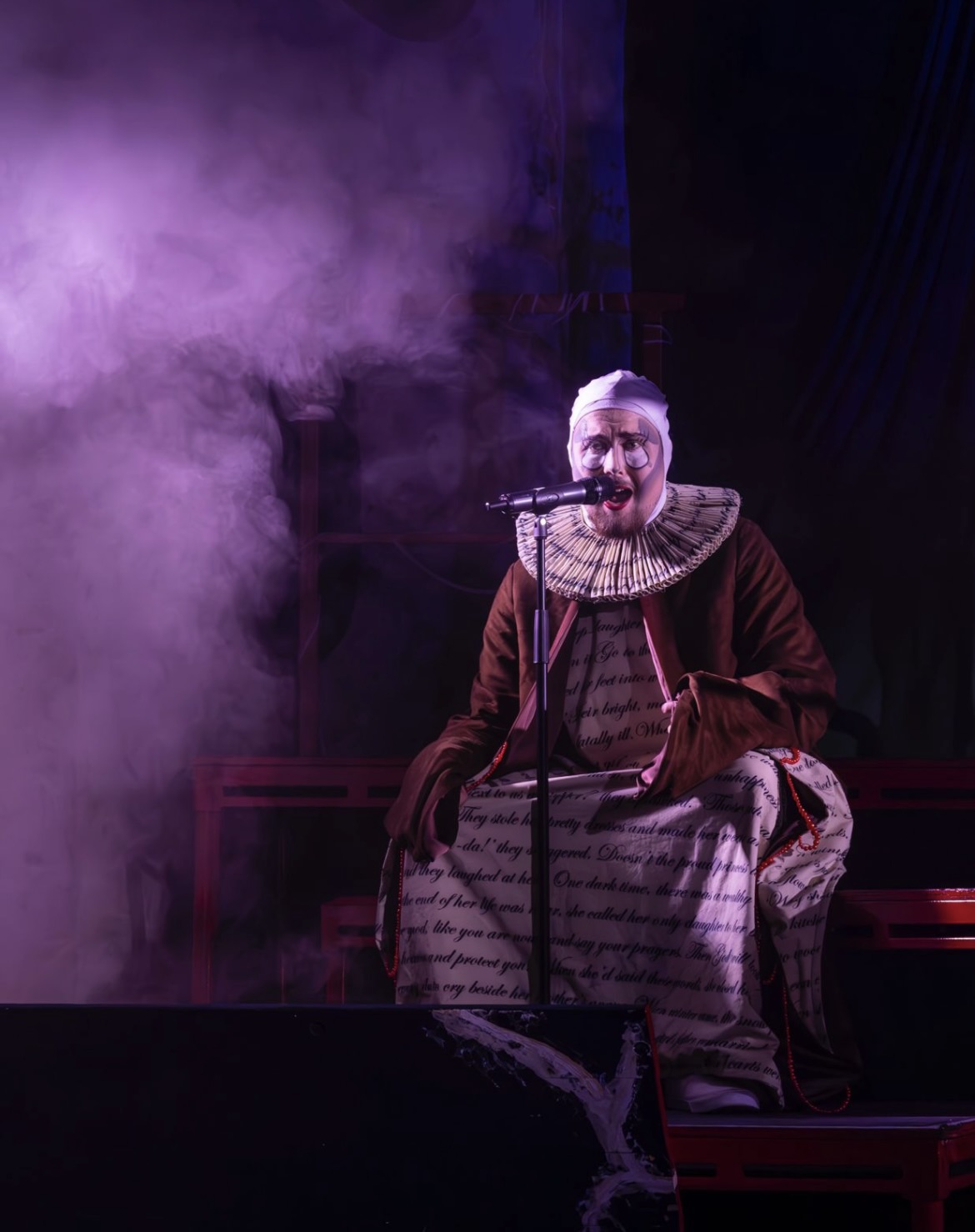 A performer seated on a stage wearing a large ruff collar and a costume that appears to be crafted from fabric with text printed on it. The background features a fog effect and dim purple lighting.