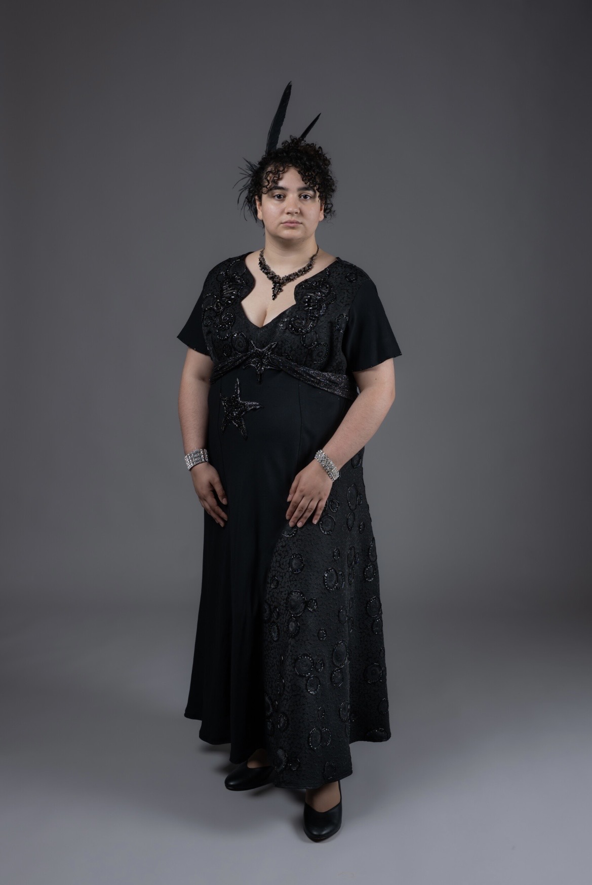A person wearing a black, floor-length dress adorned with intricate beading stands against a plain grey background. The outfit is complemented by decorative accessories, including a headpiece with feathers and jewellery.