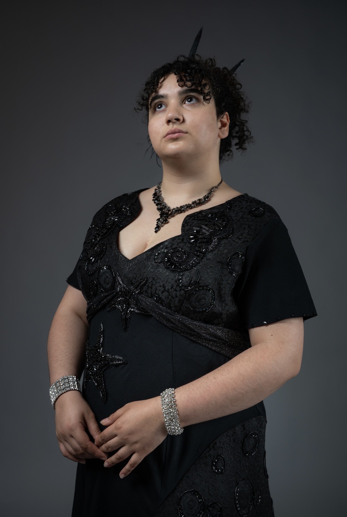 A young person wearing an elegant black dress adorned with sequins stands against a dark background. They have curly hair and are accessorised with a necklace and bracelets, with their hands clasped in front of them.