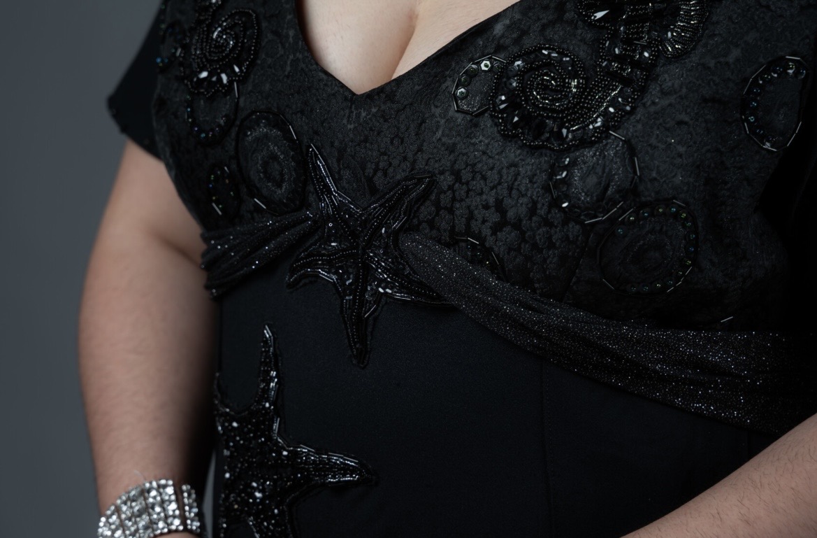 A close-up of a woman wearing a black dress adorned with intricate sequined and beaded designs. The neckline is deep, and she is accessorised with a silver bracelet on her wrist, with a plain dark background.