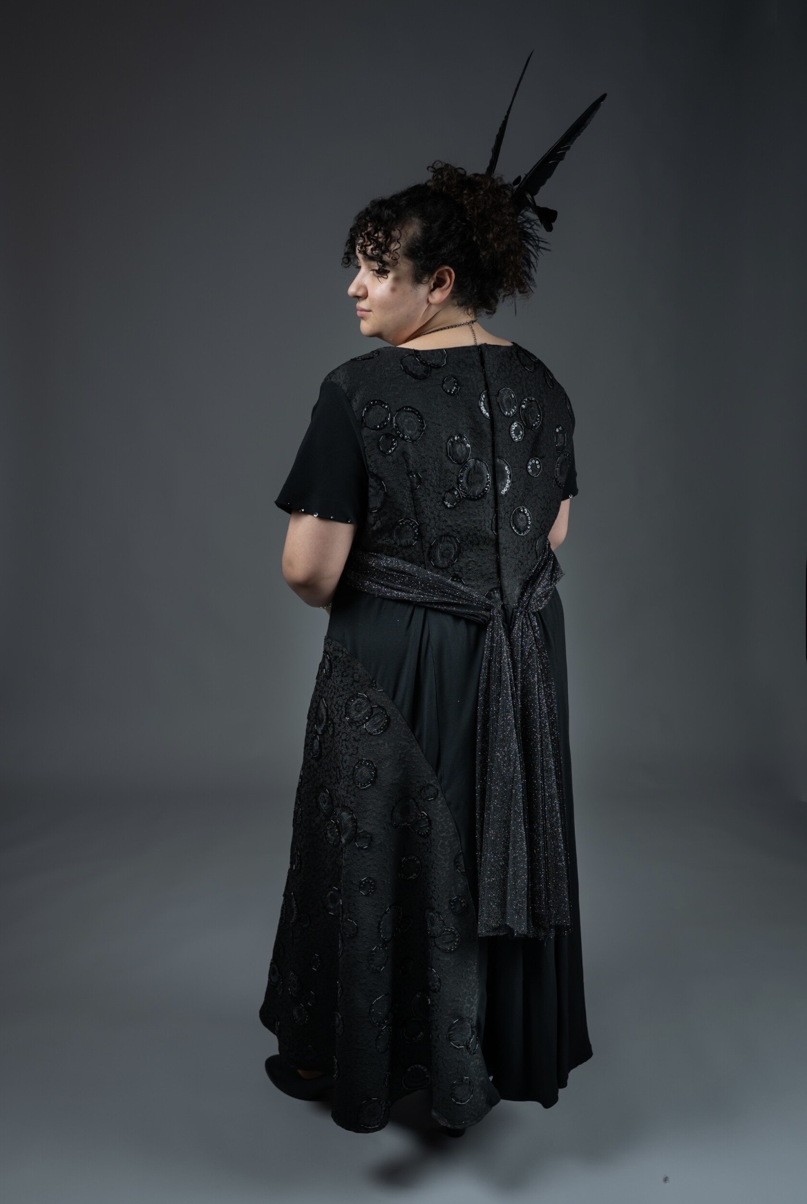 A woman with curly hair wearing a black dress with intricate details stands with her back to the camera. The dress features a sash tied at the waist and is complemented by a decorative headpiece with feathers, set against a grey background.