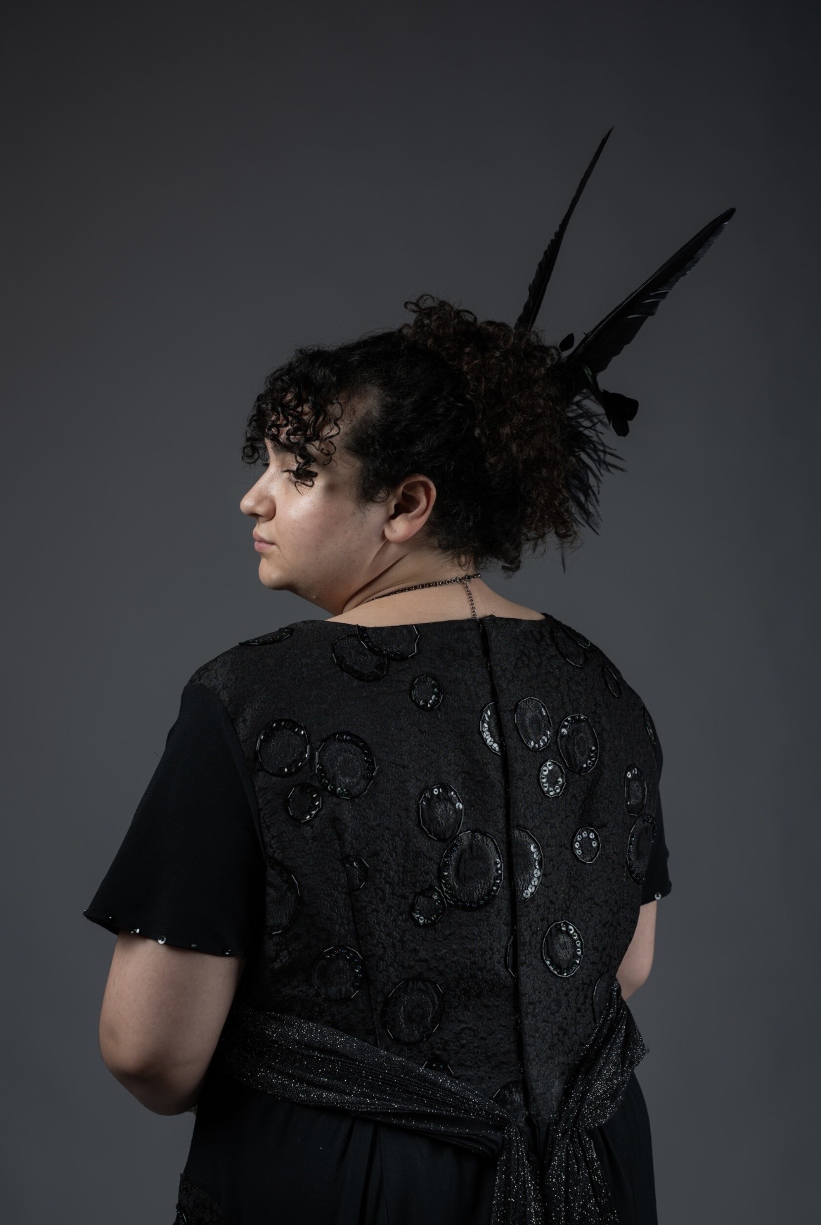 A young person with curly hair styled up, wearing a black dress with decorative embellishments, stands with their back to the camera. A feathered accessory is positioned at the top of their head, against a dark grey background.