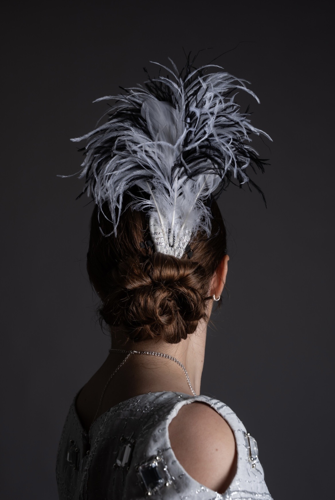 A woman with auburn hair styled in a low bun is seen from behind, wearing a silver gown. A decorative headpiece featuring white and black feathers is positioned atop her head against a dark backdrop.