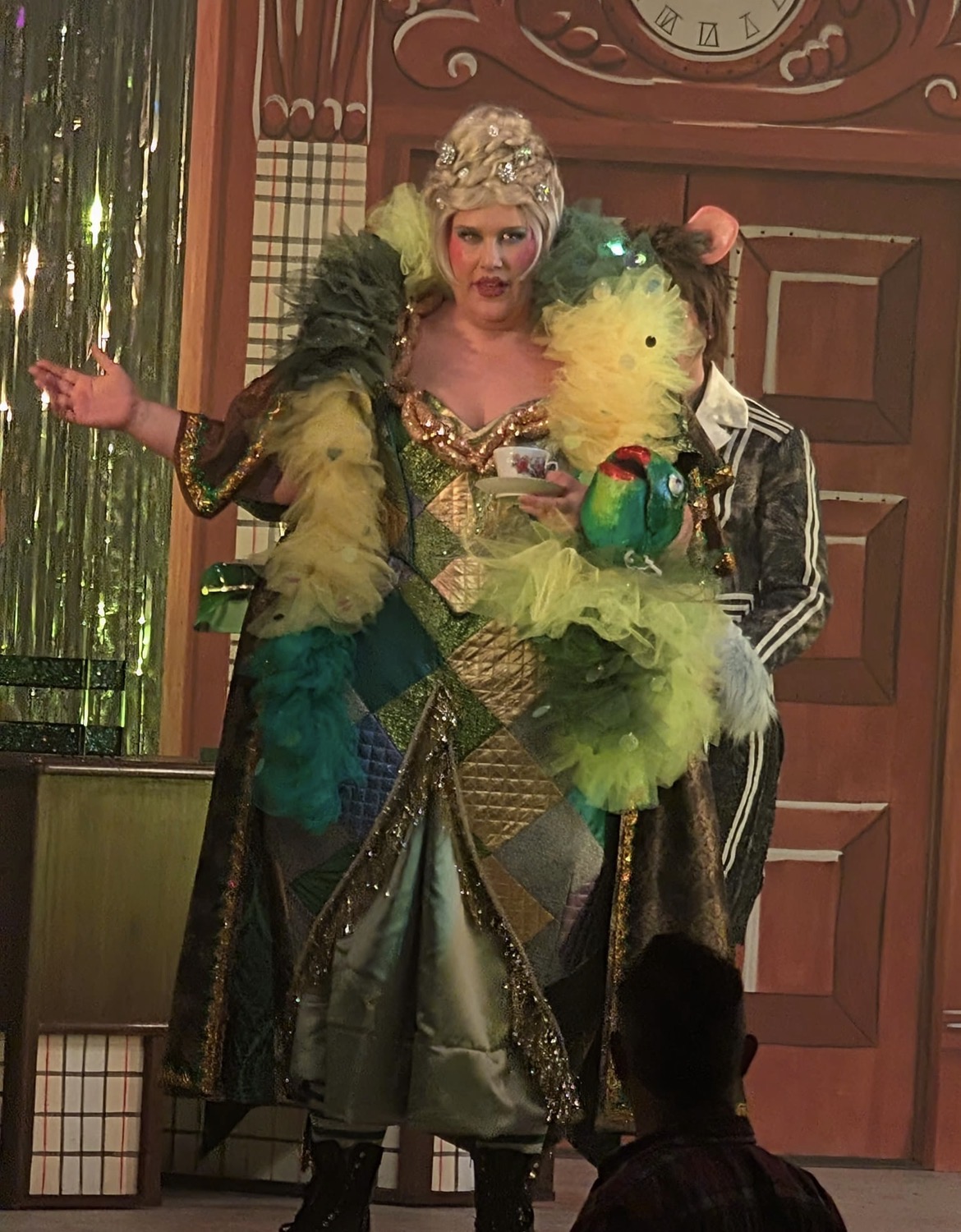 A performer wearing a glittering gold dress with a patchwork design, adorned with colourful feathers, stands on stage. They are holding a cup in one hand and gesturing with the other, while another person in a striped suit stands behind them.