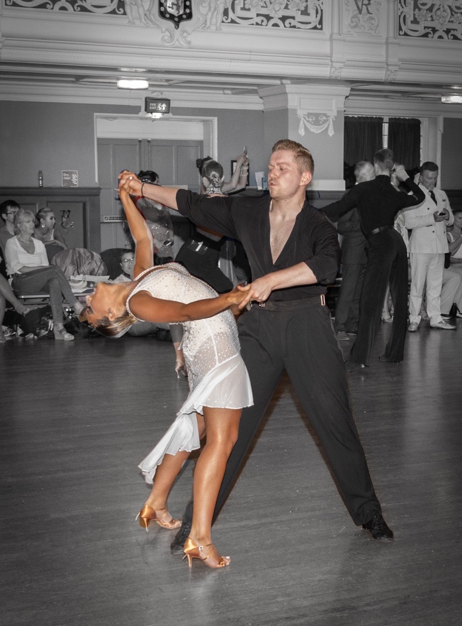 Ballroom dancing couple performing in ornate hall, woman in white dress dipped backwards, man in black suit, other dancers in background in black and white..