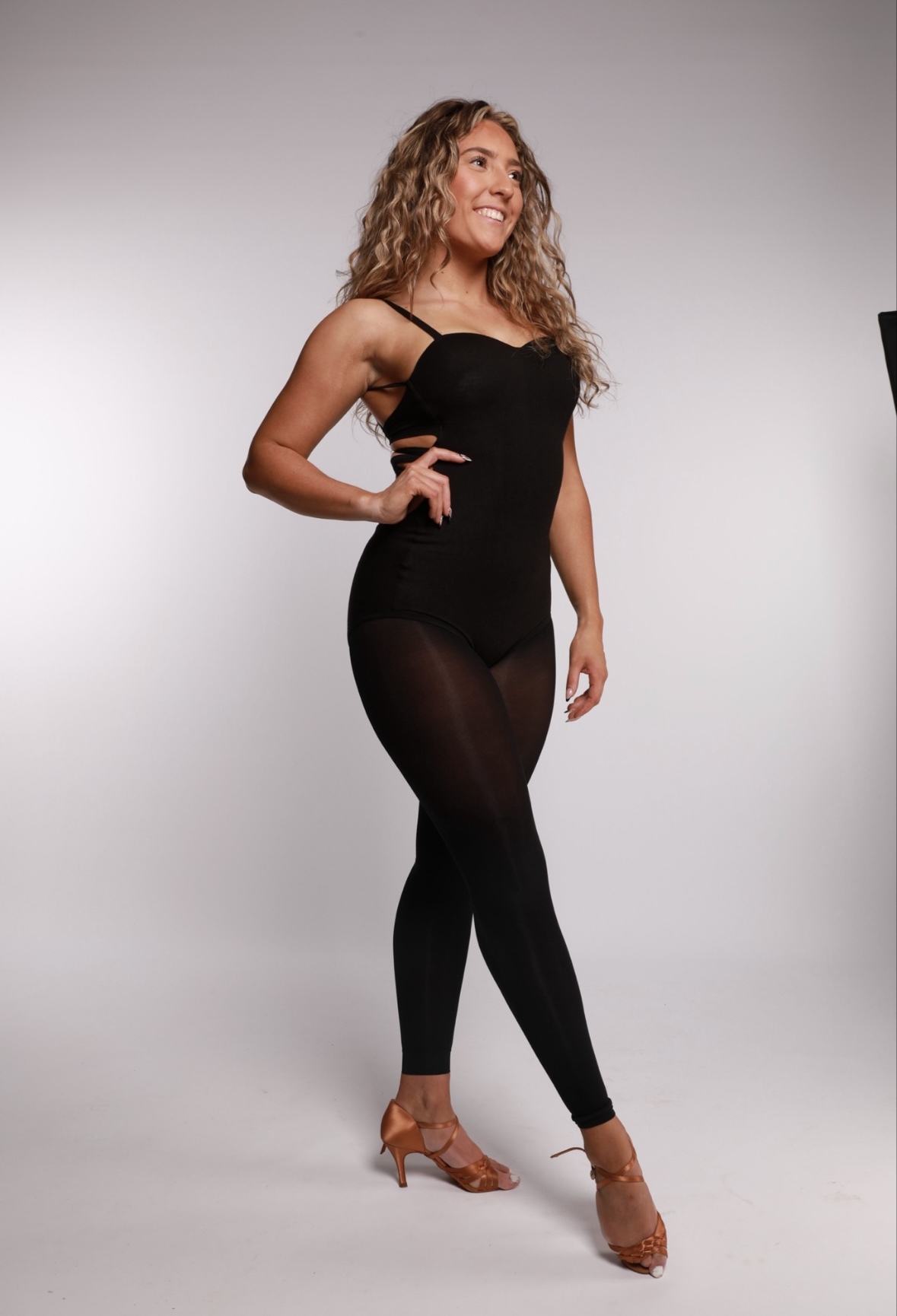 Woman with wavy blonde hair wearing black fitted dress with mesh panel and brown strappy heels, posing against grey background.