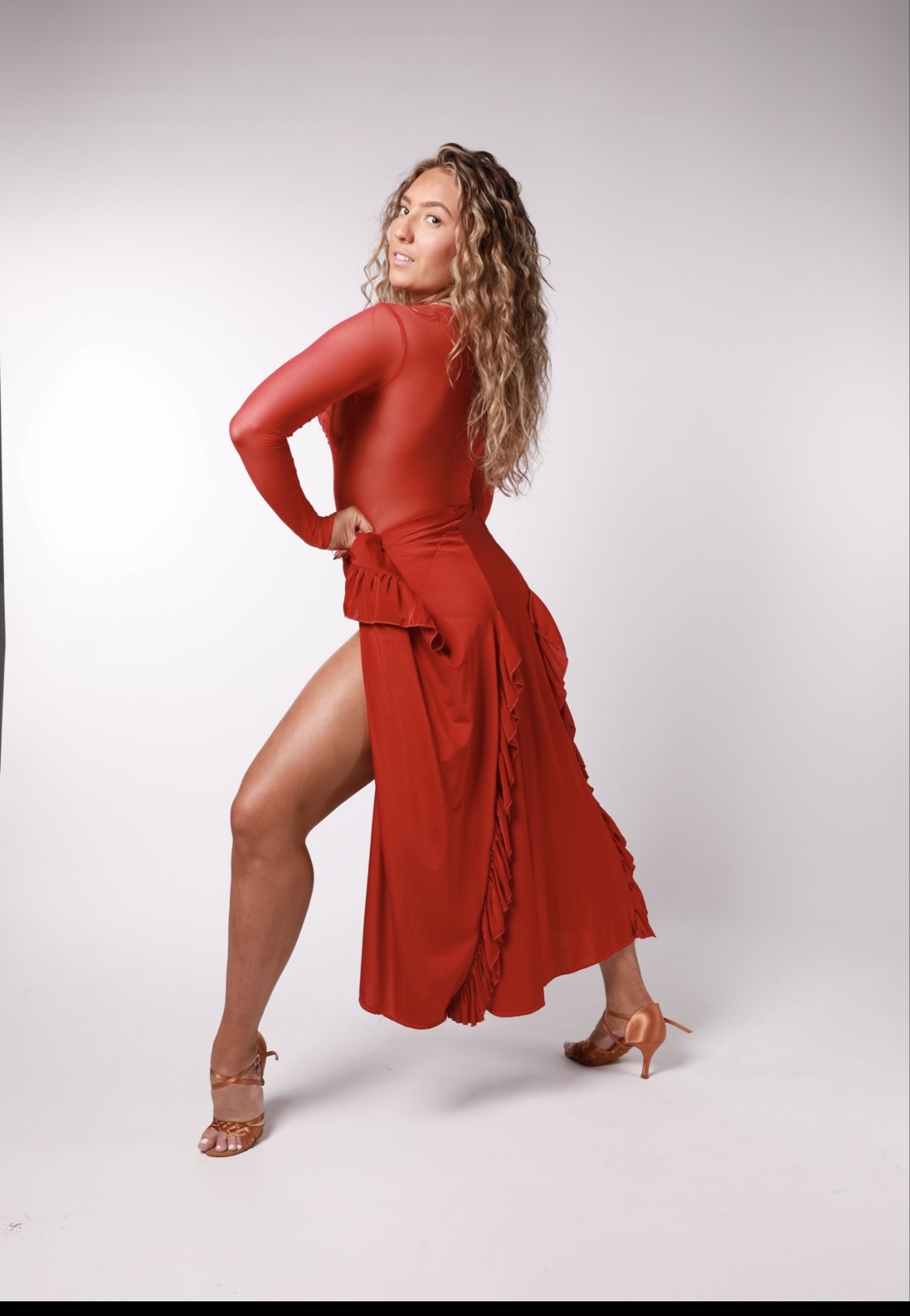 Woman with long wavy blonde hair wearing red long-sleeved top and ruffled skirt with high slit, brown strappy heels, grey background.
