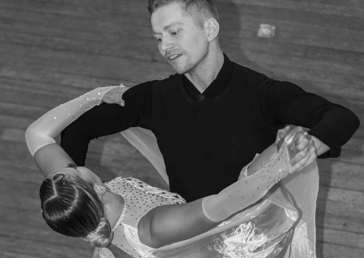 Man in black top dancing with woman in light-coloured dress, captured mid-movement against brick wall background. Black and white image.