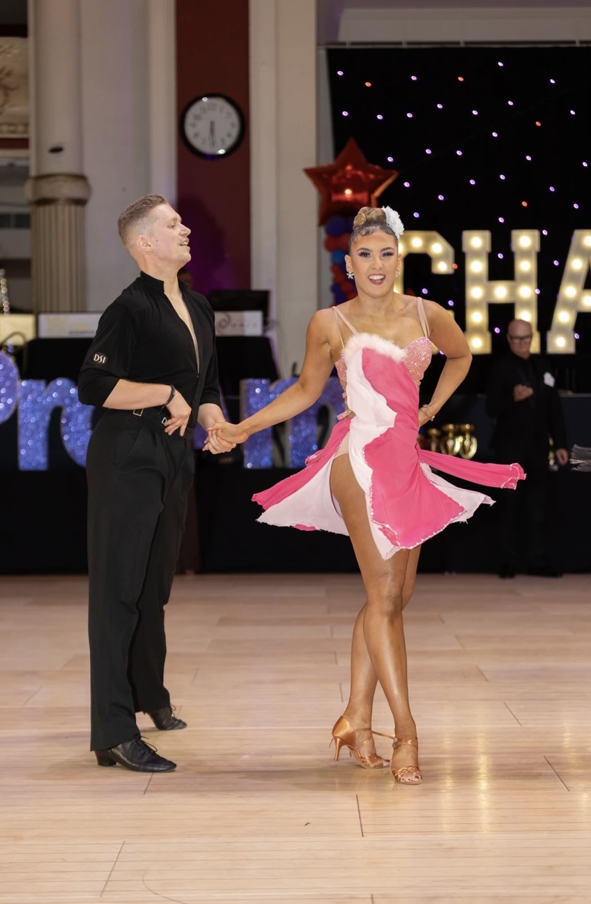 Two ballroom dancers performing on wooden floor, man in black outfit, woman in pink and white dress with sequins and fringe detail.