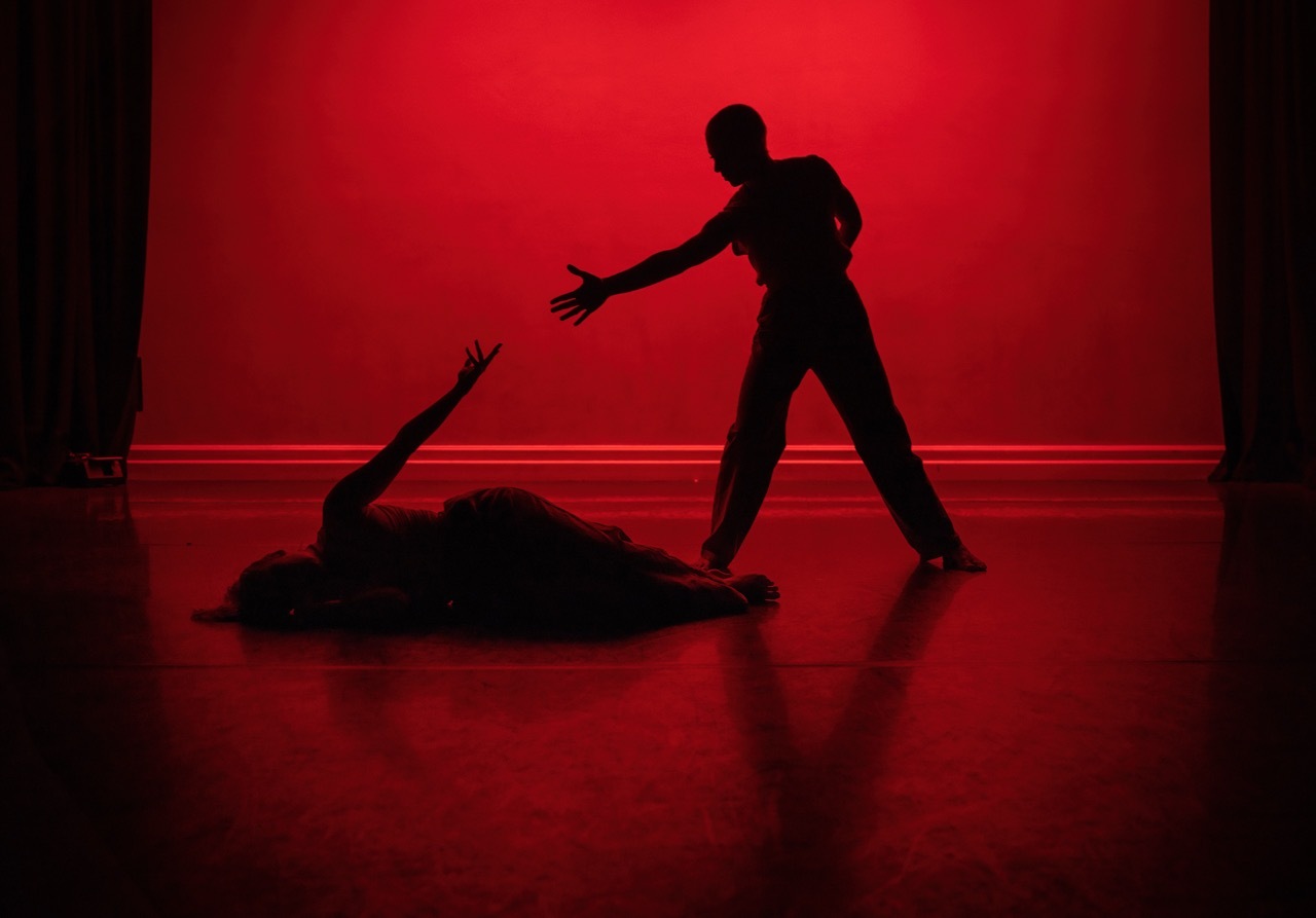 Two dark silhouettes against bright red backdrop - one figure standing with outstretched arm, another lying on reflective floor.