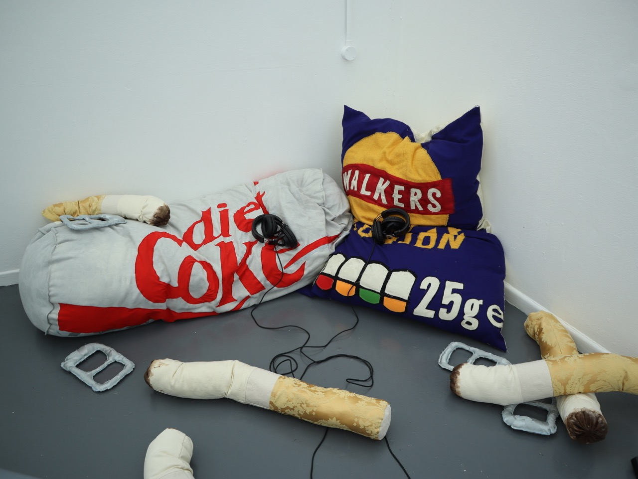 Two oversized bean bags styled as branded packaging, one resembling a Diet Coke can and the other a Walkers crisps bag, are placed on a grey floor. Several fabric sculptures resembling food items are scattered around them.