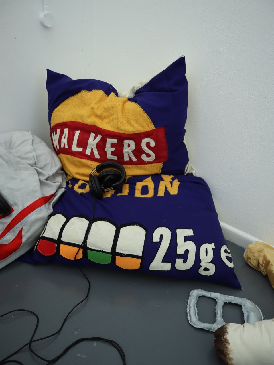 A large cushion designed to resemble a Walkers Crisps packet, featuring the word 'WALKERS' in red and yellow. The cushion is placed on a grey floor next to a grey blanket and a pair of headphones.