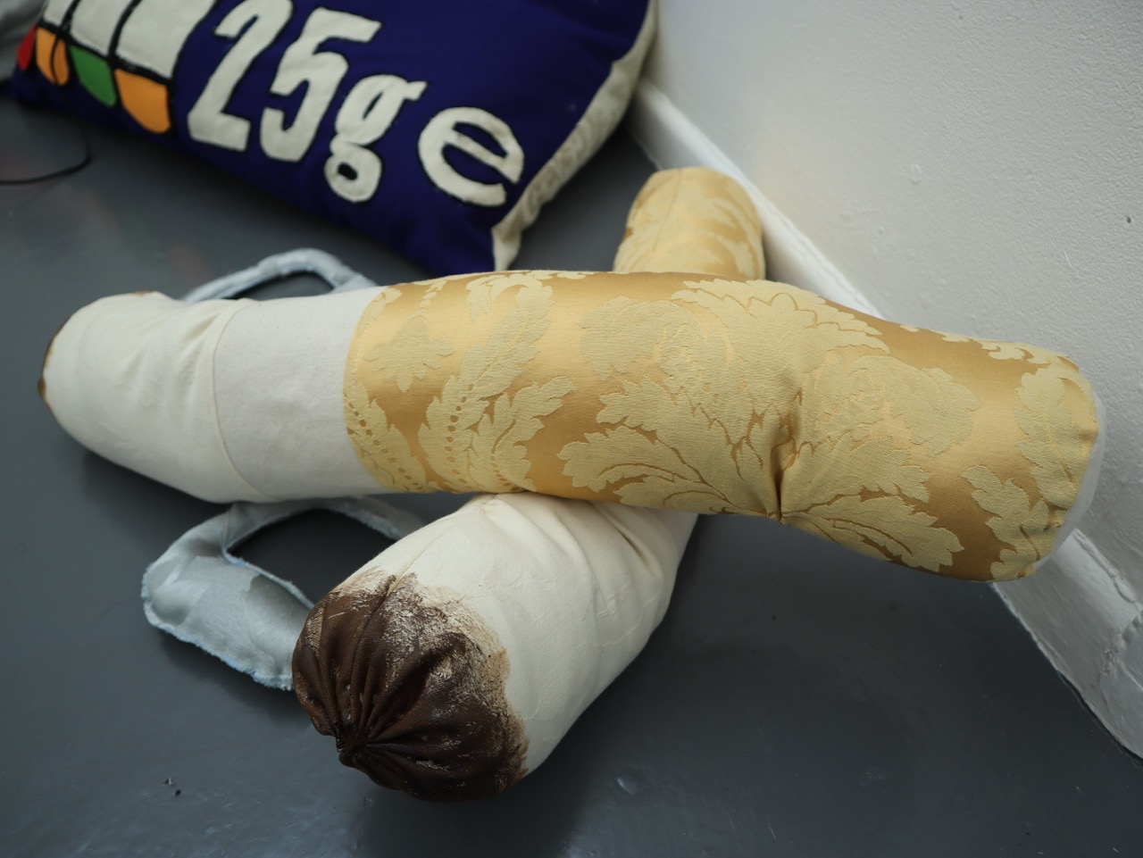 Two decorative bolster cushions are stacked on a grey floor. The top cushion features a gold patterned fabric, while the bottom cushion has a white fabric covering with a brown end. A colourful fabric with numbers is visible in the background.