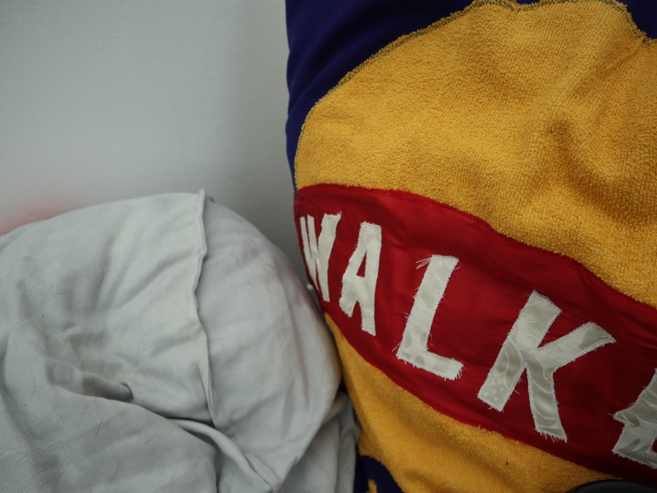 A corner of a bed with a crumpled white pillow and a close-up of a bright yellow and purple blanket featuring the word "WALKER" in bold white letters and red. The background is plain and minimalistic.