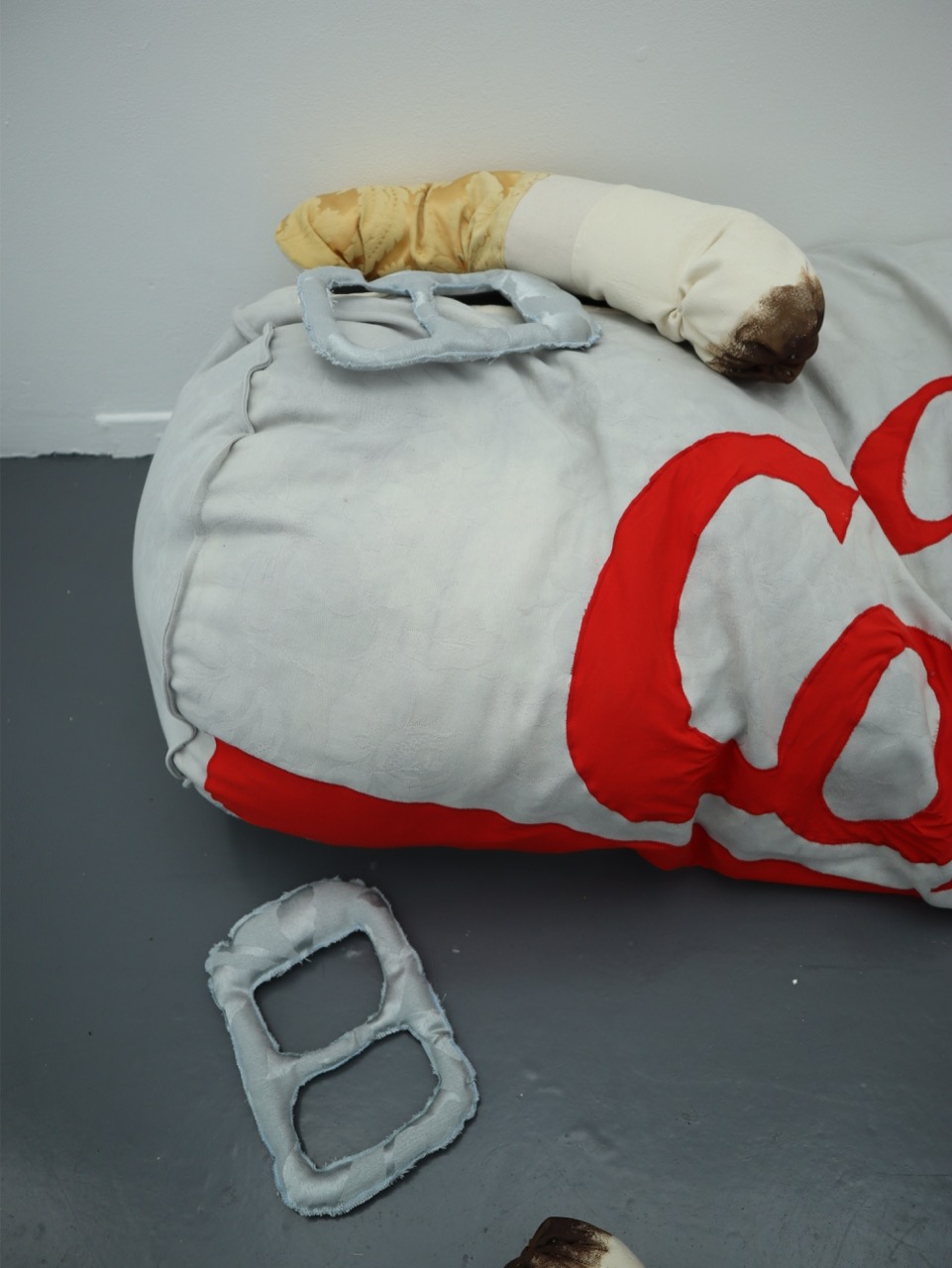A large, soft cushion in the shape of a food item features grey and red colours. Nearby, two grey handles resembling those of a sandwich are placed on the floor, positioned beside a light brown curved object.