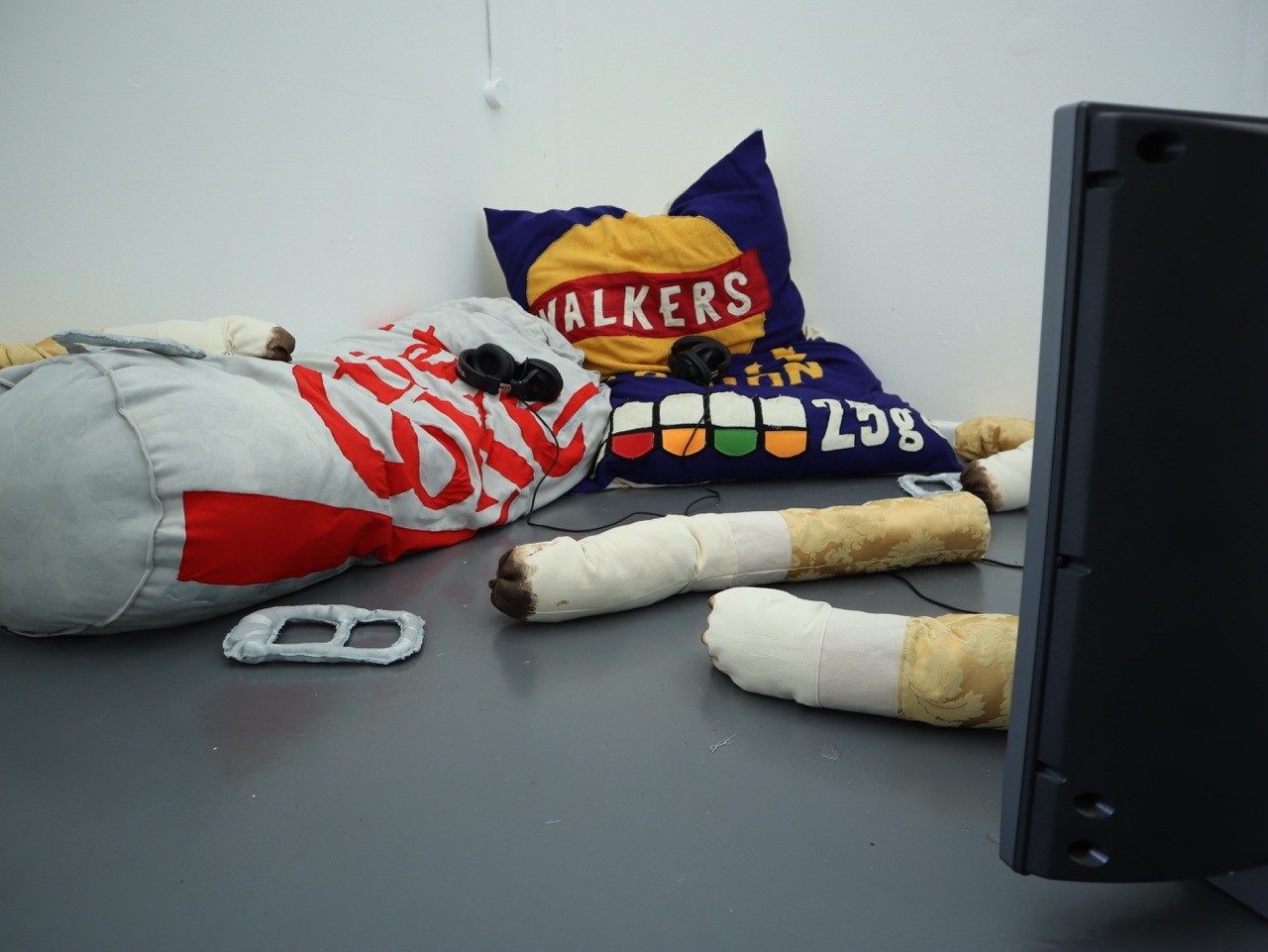 A cozy seating area with a large, soft pillow designed to resemble a bag of Walkers crisps, accompanied by a colourful cushion. Nearby, a monitor displays a simple screen. A pair of plush arms with bandages is draped over the side.