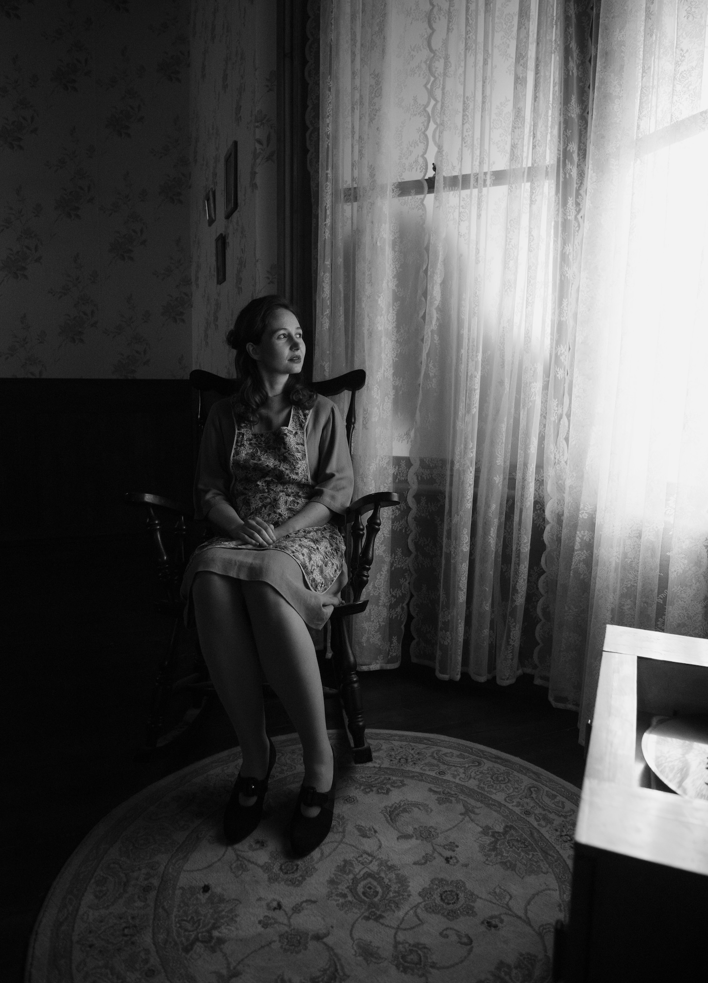 Black and white still from "Lullaby for the Lost". A woman sits in a rocking chair and looks to the right out of two tall windows, covered with net curtains.