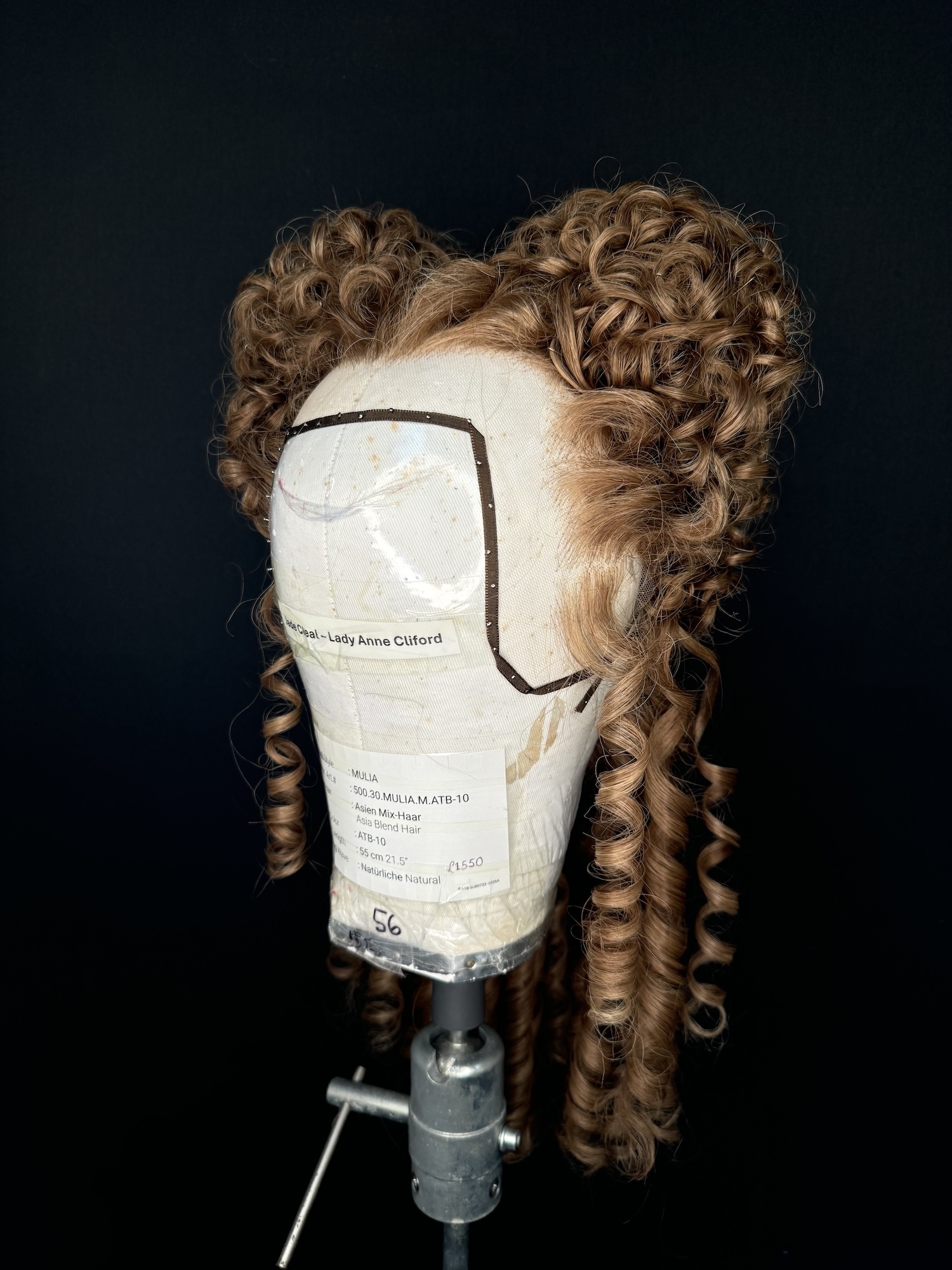 A brown-haired curly wig in a historical style on display on a mannequin head against a black background.