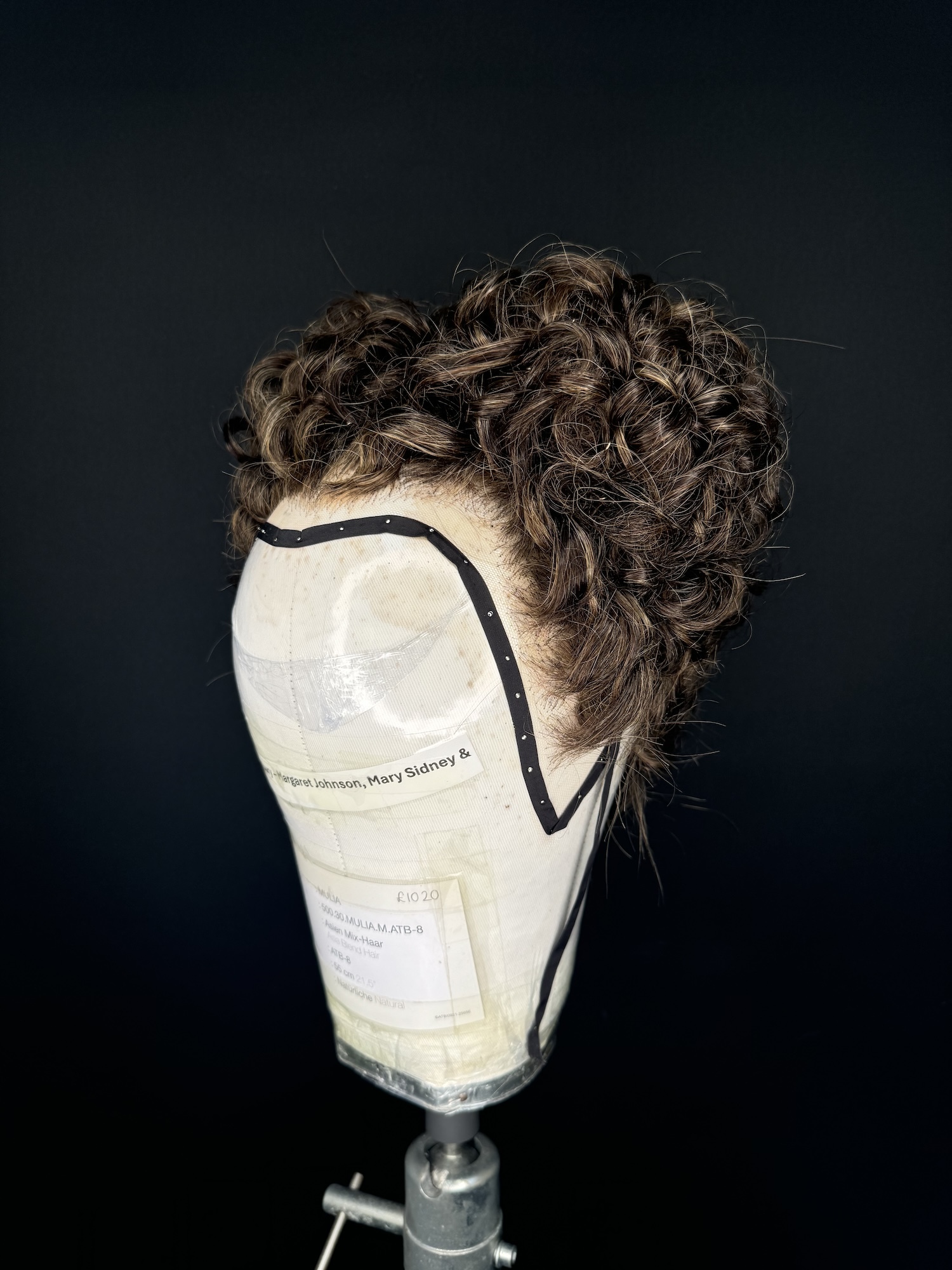 A dark-haired wig in a historical style on display on a mannequin head against a black background.