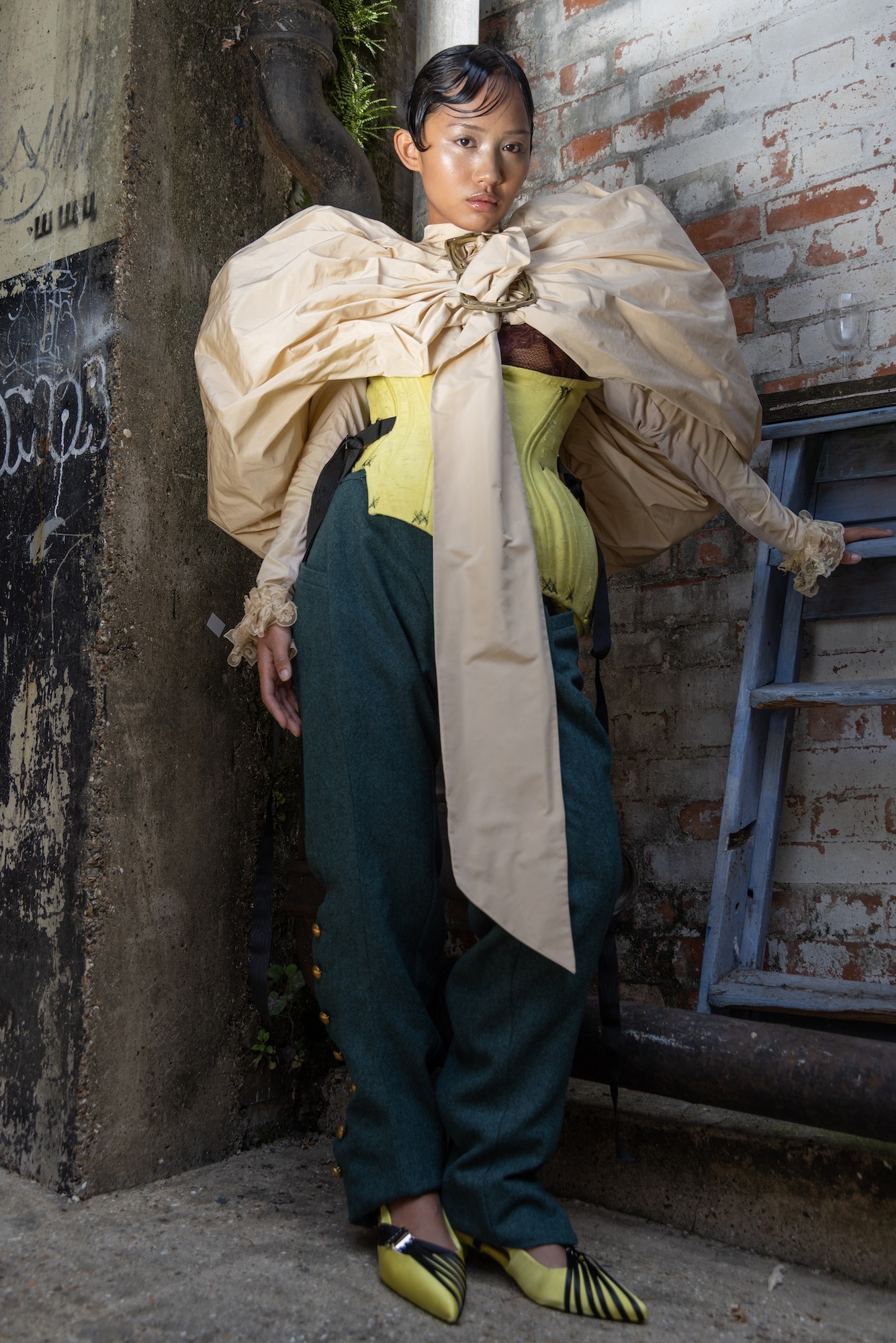 Model wearing a yellow, irregular-shaped top, a pair of dark blue trousers and a pale gold puffy coat/cape.