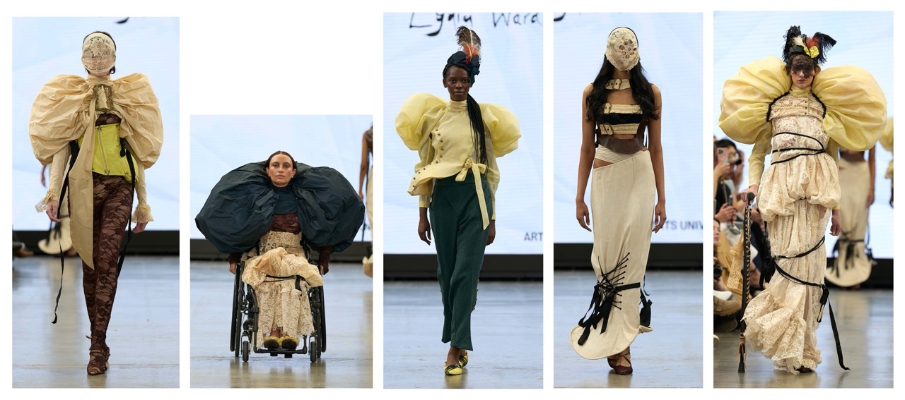 Five fashion models on white runway wearing avant-garde designs with large blonde hair pieces, earth tones, and dramatic silhouettes including oversized collars and layered fabrics.