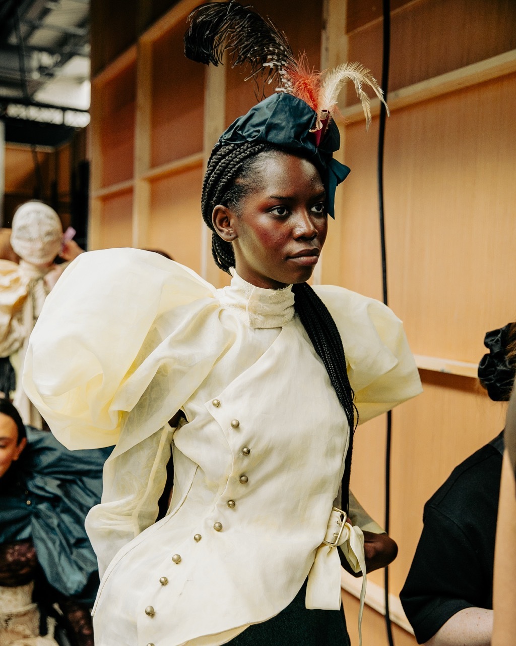 Black woman in white Victorian-style dress with puffy sleeves and buttons, ornate headpiece with feathers, in period room setting.