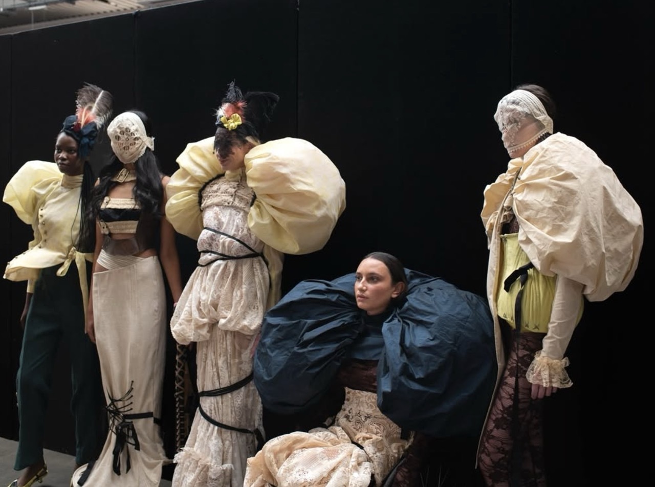 Five models on stage in period costume; centre figure in dark blue dress kneels, others wear cream and white garments with puffy sleeves.