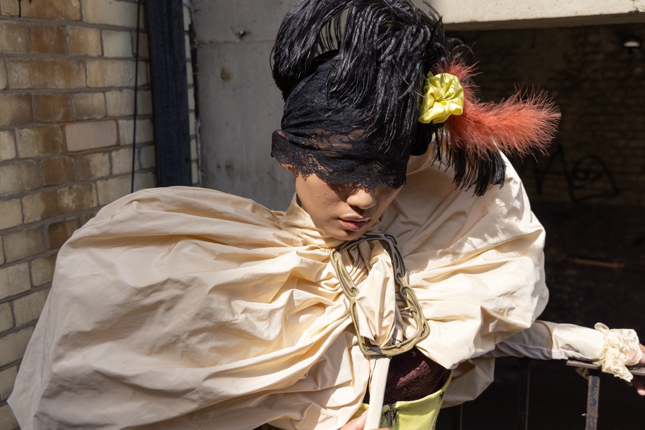 Person in period costume with black tricorn hat adorned with yellow and red feathers, wearing cream-coloured flowing shirt with rope belt.