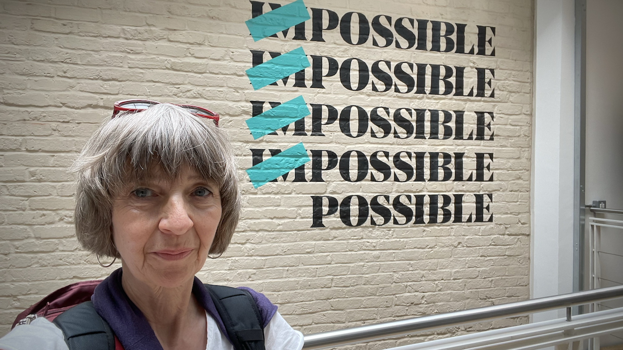 Woman with grey bob haircut stands before white brick wall with "IMPOSSIBLE" repeated five times, teal checkmarks over "IM" prefixes.