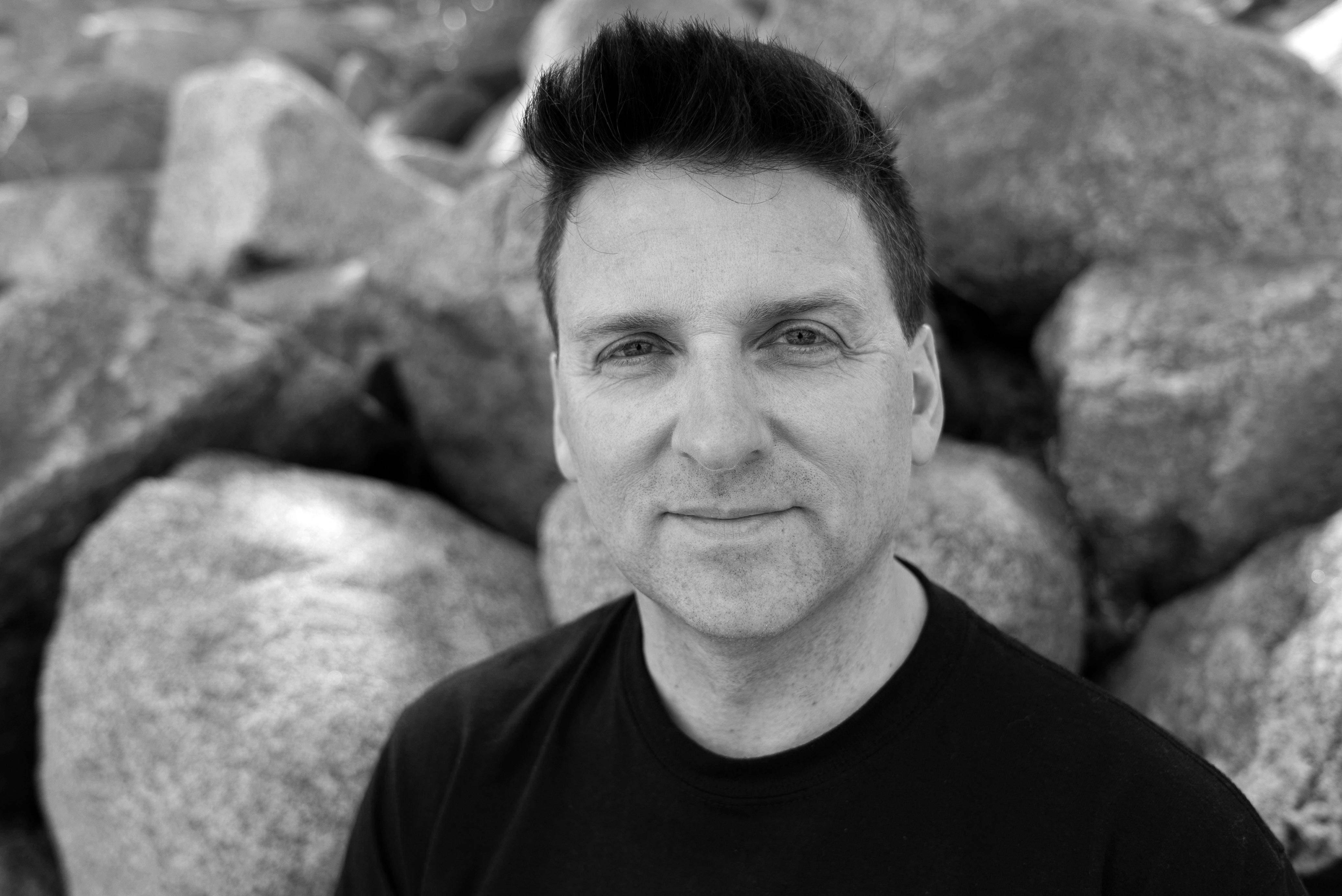 Black and white portrait of man with dark hair wearing black shirt, stone wall background.