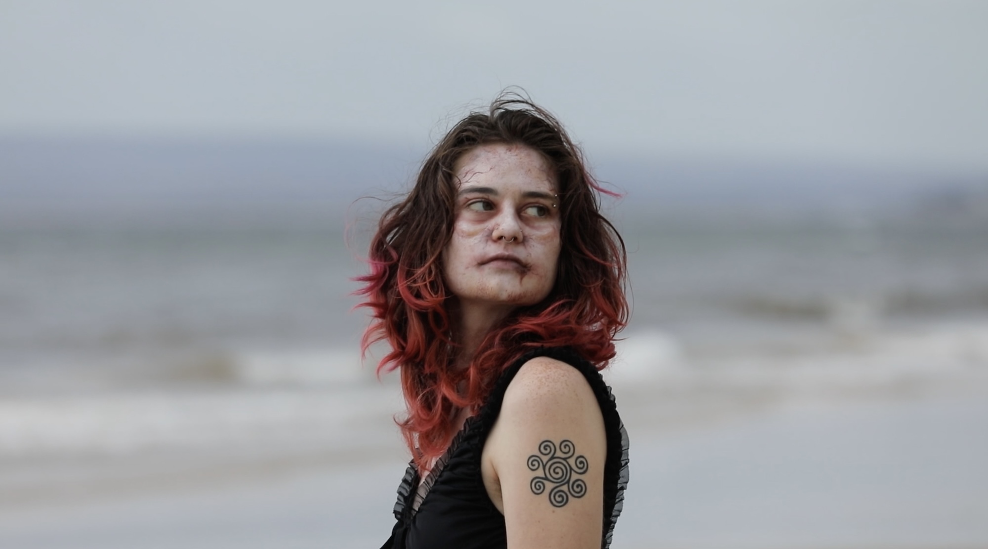 A model wearing horror/corpse-style make-up with red hear and a swirly tattoo on their arm, looking off to the right. The sea is blurred in the background.