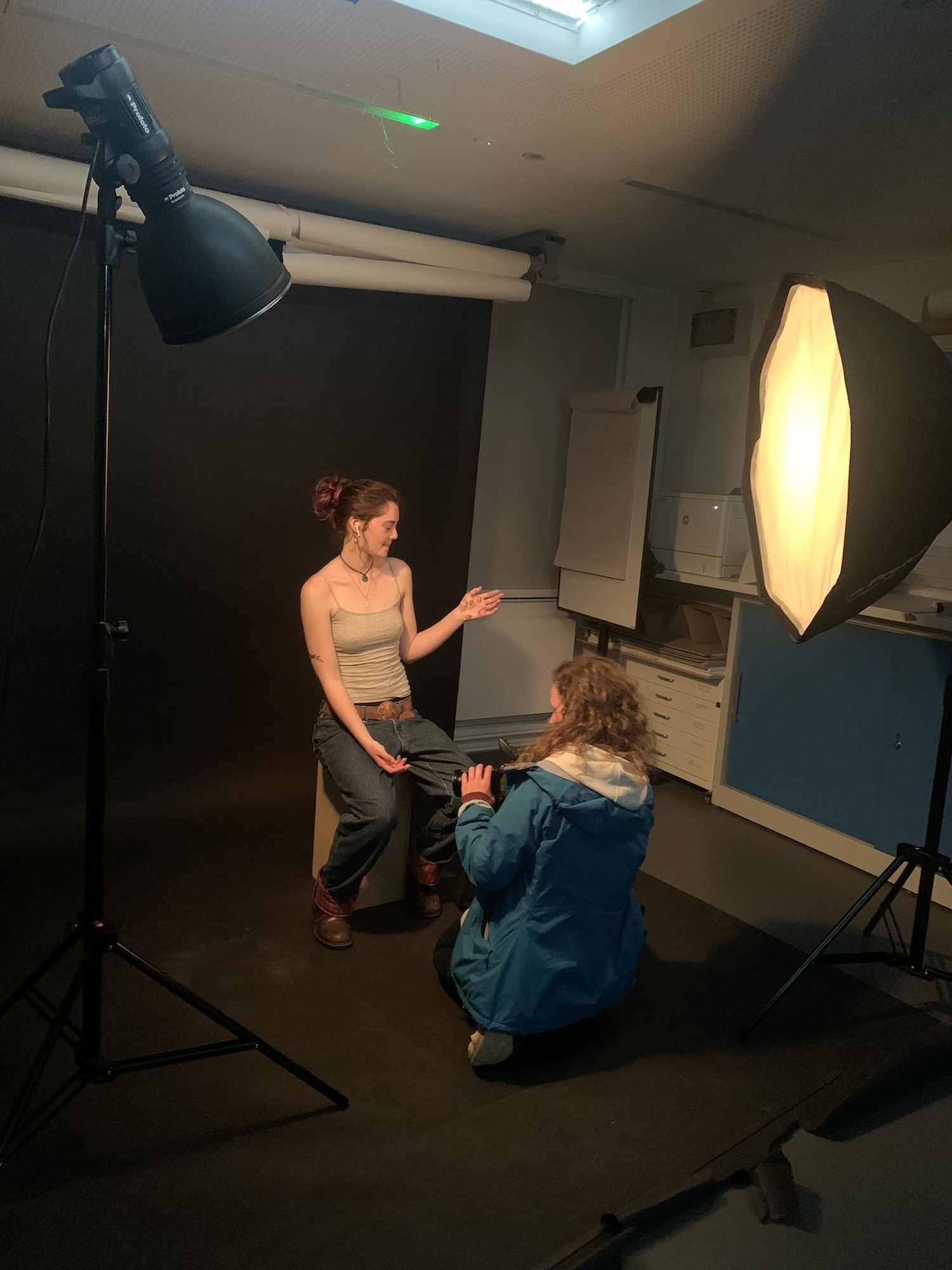 Two Foundation Diploma in Art, Design and Media students in a studio space with a different light either side of them. One student is sitting on a box, the other is knelt in front of them preparing to apply make-up.