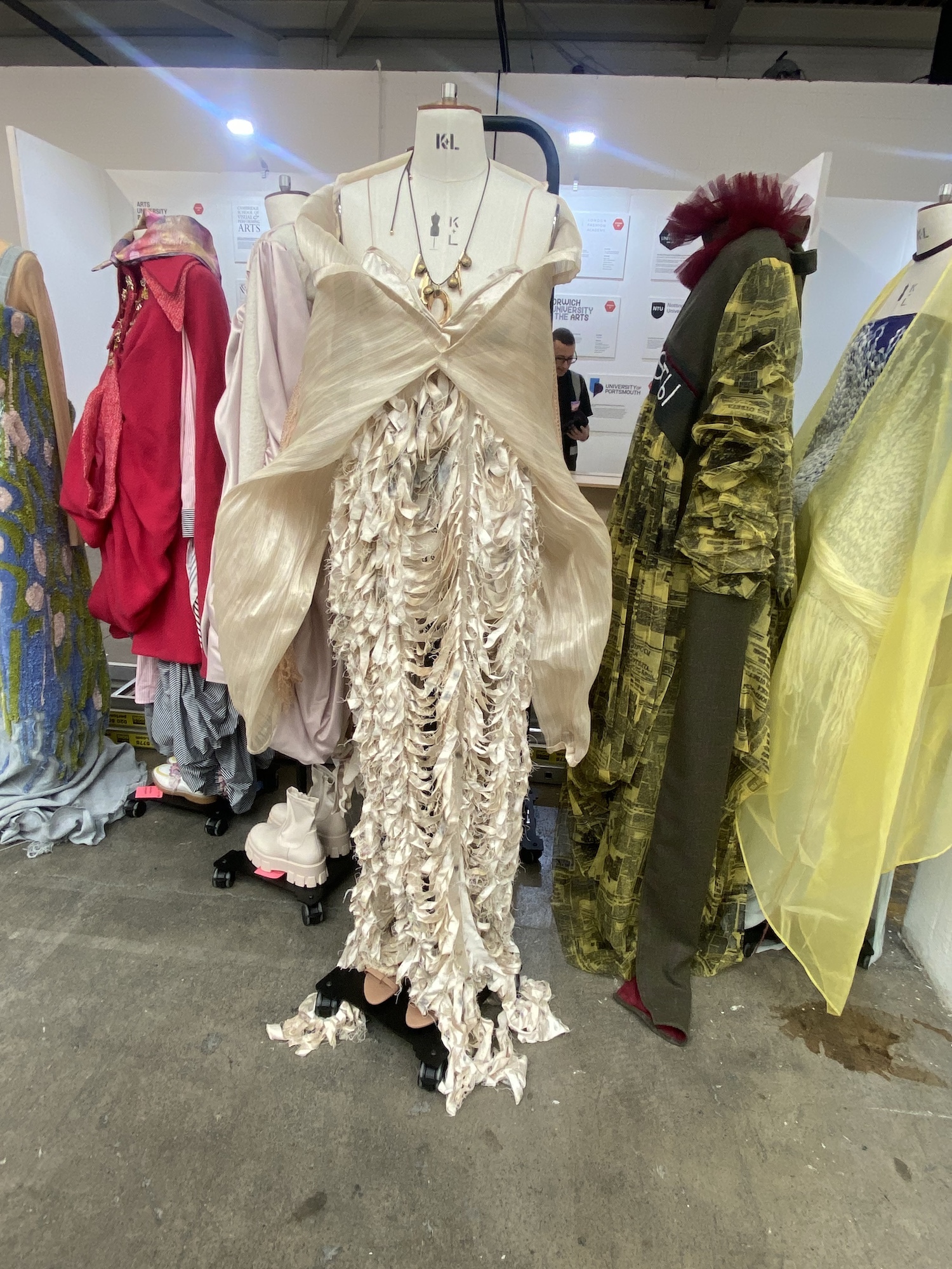 A flowing white dress and shawl on a mannequin, with several other garments around it, at Graduate Fashion Week 2024.