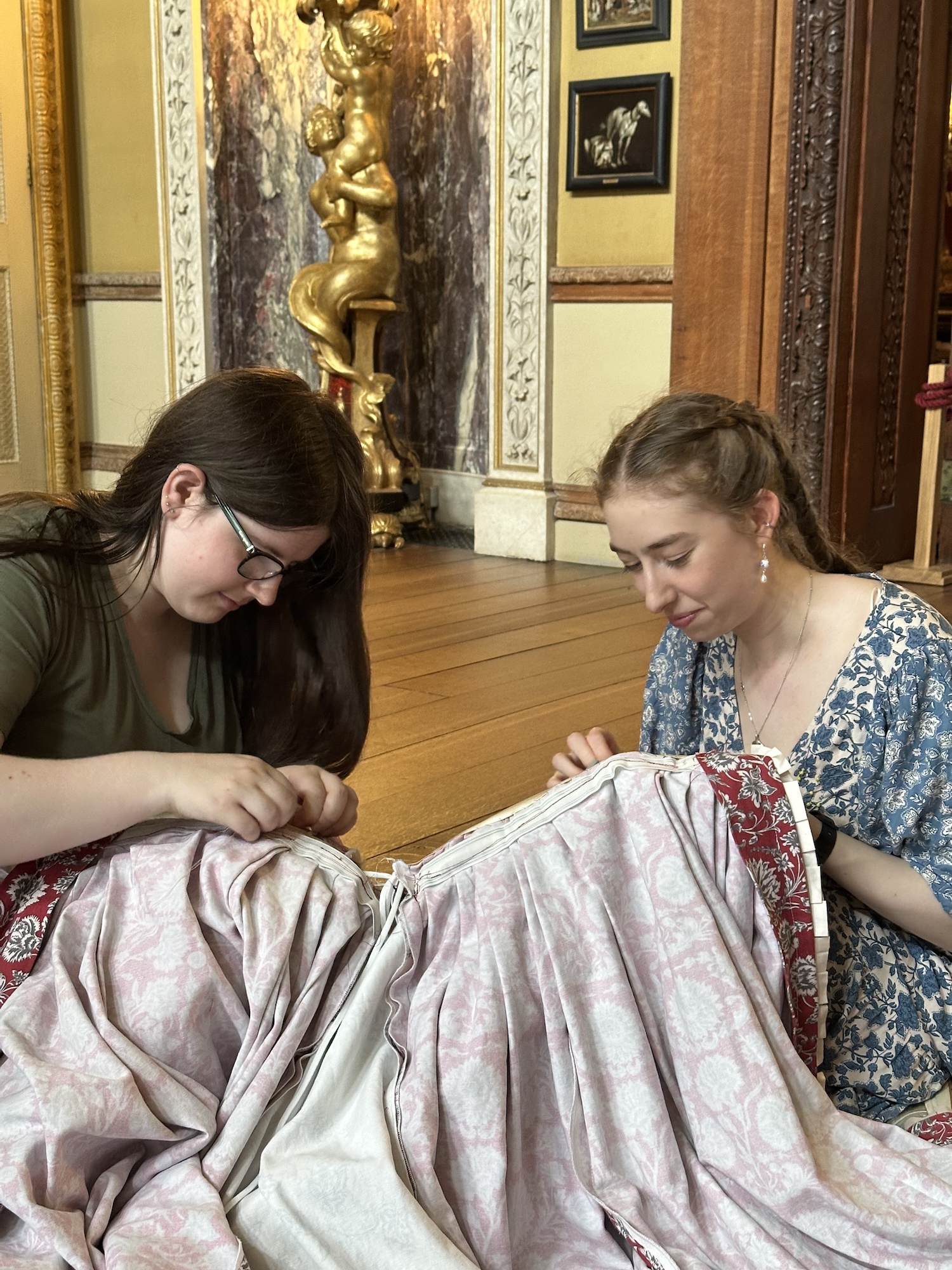 Two MA Historical Costume students are sewing fabric together while seated on the floor in a grand room with wooden flooring and ornate decorations. The background features a golden statue and detailed wall designs.
