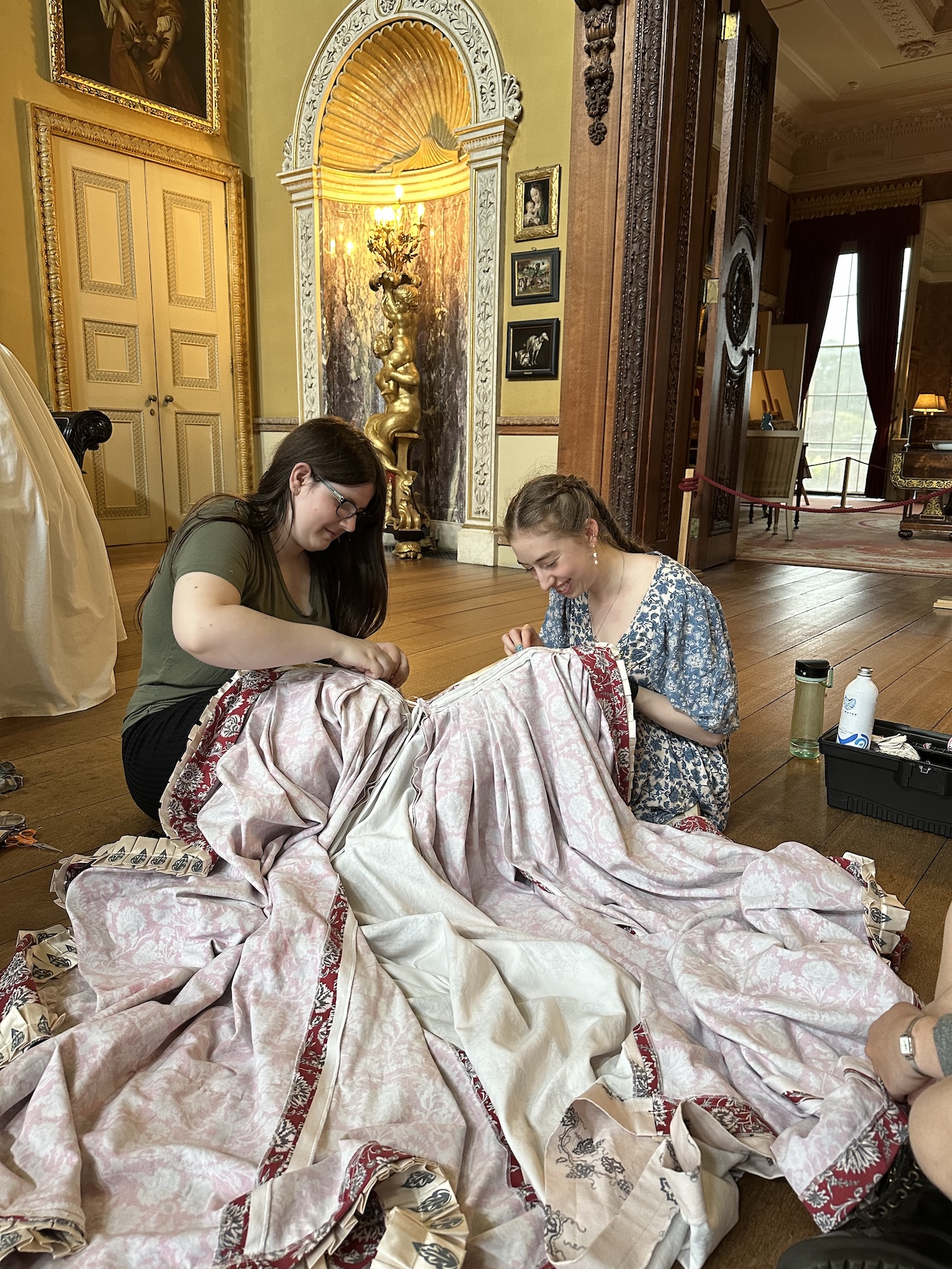 Two MA Historical Costume students are sitting on the wooden floor of a grand room, working on a large piece of patterned fabric. One woman has long brown hair and wears a green top, while the other has braided hair and a blue dress. The room features ornate decorations and large windows.
