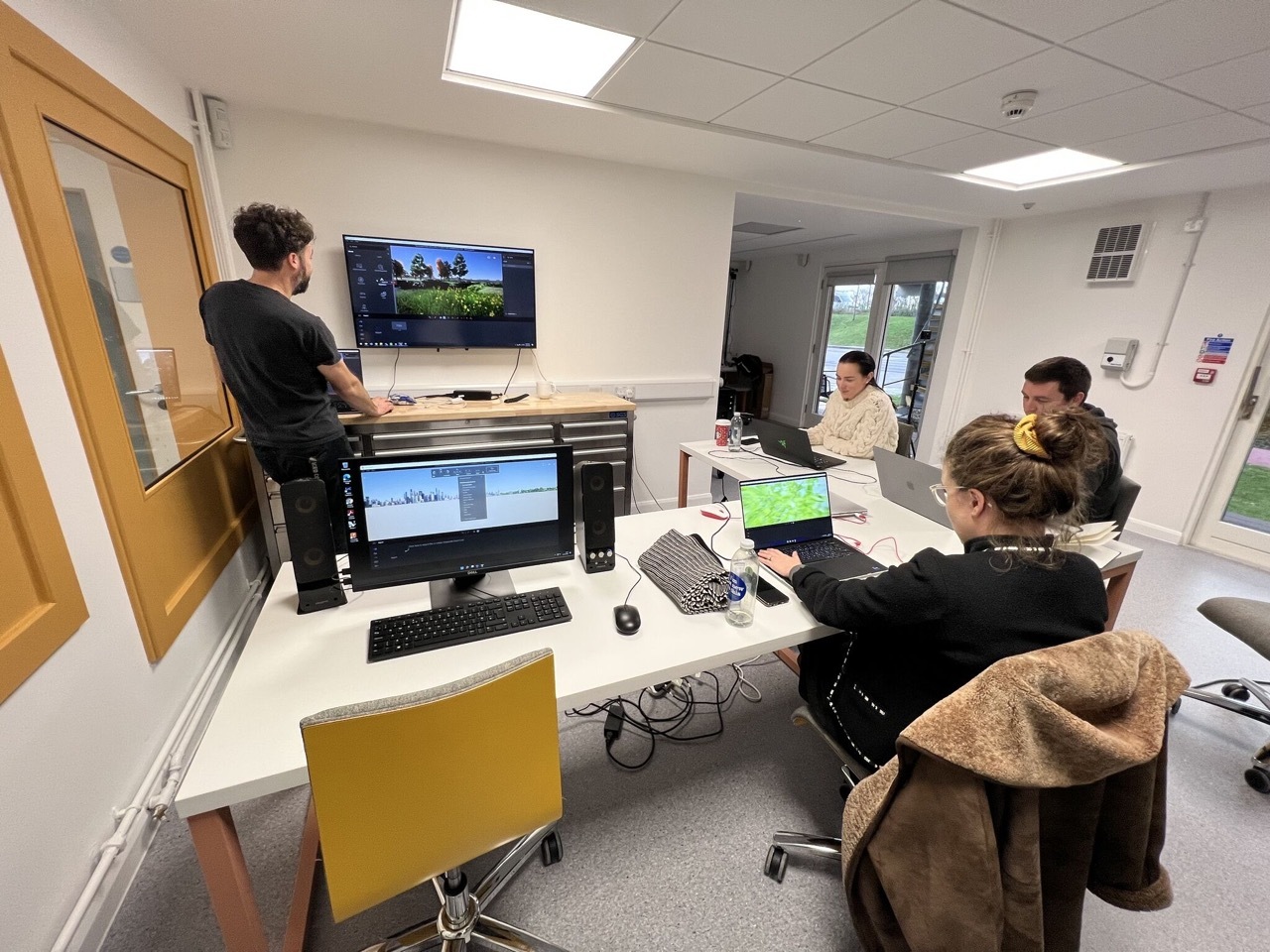 Students working in a bright studio with multiple computers and screens, yellow chairs and frames on the left wall.