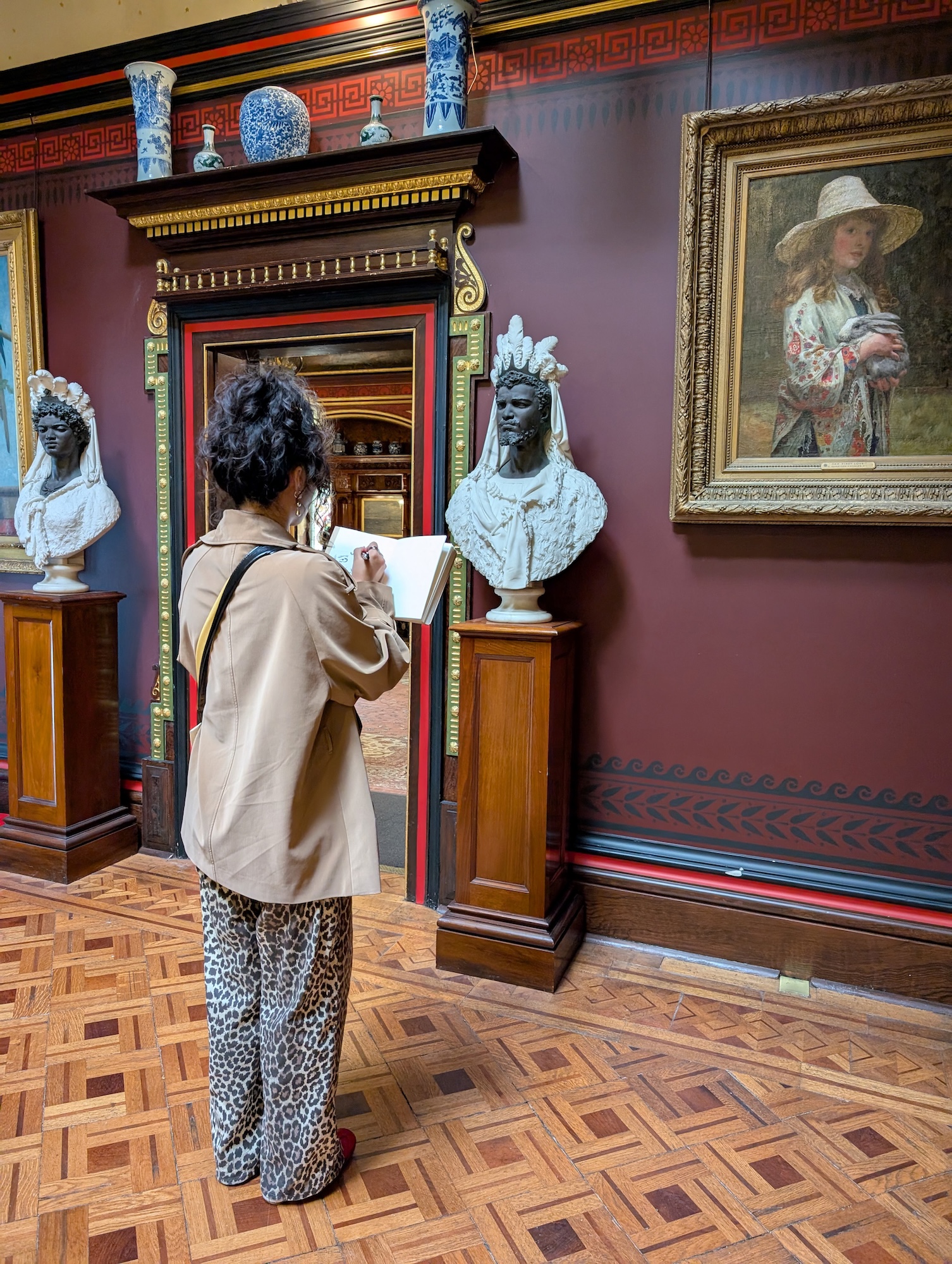 A woman with curly hair wearing a beige jacket and patterned trousers stands in a museum gallery. She is sketching from two white busts displayed on pedestals, with a painting of a girl in a hat hanging on the wall to her right.