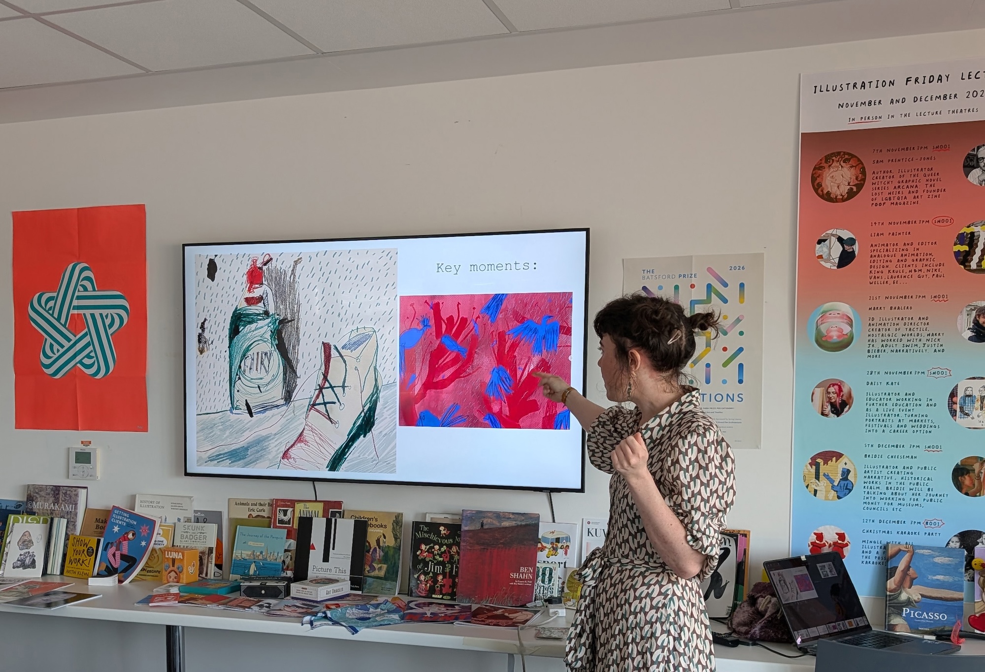 A woman wearing a patterned dress gestures towards a large screen displaying artwork during a presentation. The room features a table with books and colourful posters on the walls, including a red flag and illustrations.
