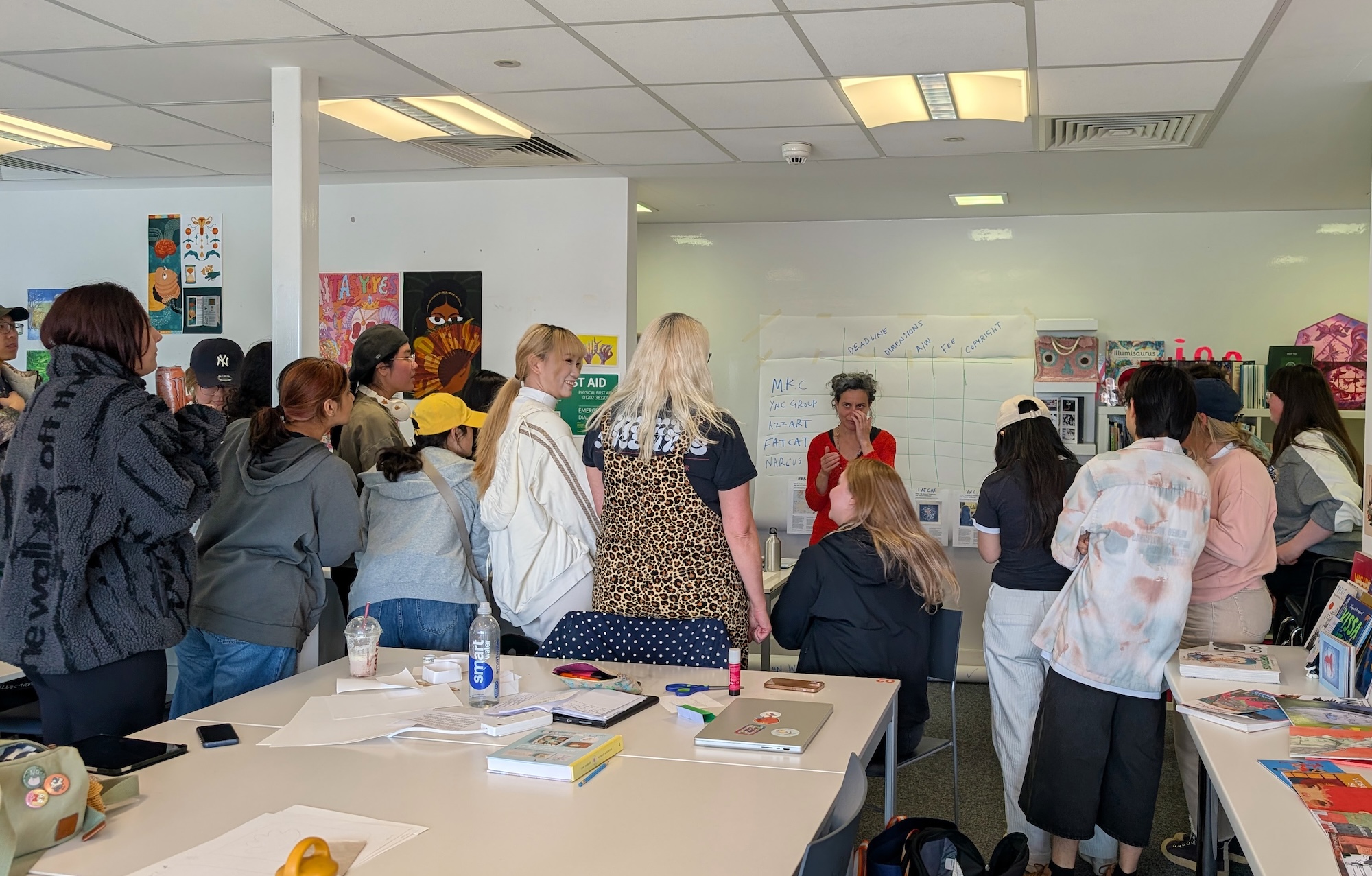 A group of students gathered in a classroom, where a woman is speaking at the front. The students are standing and sitting around various tables covered with papers and art materials. The room has bright lighting and colourful artwork on the walls.