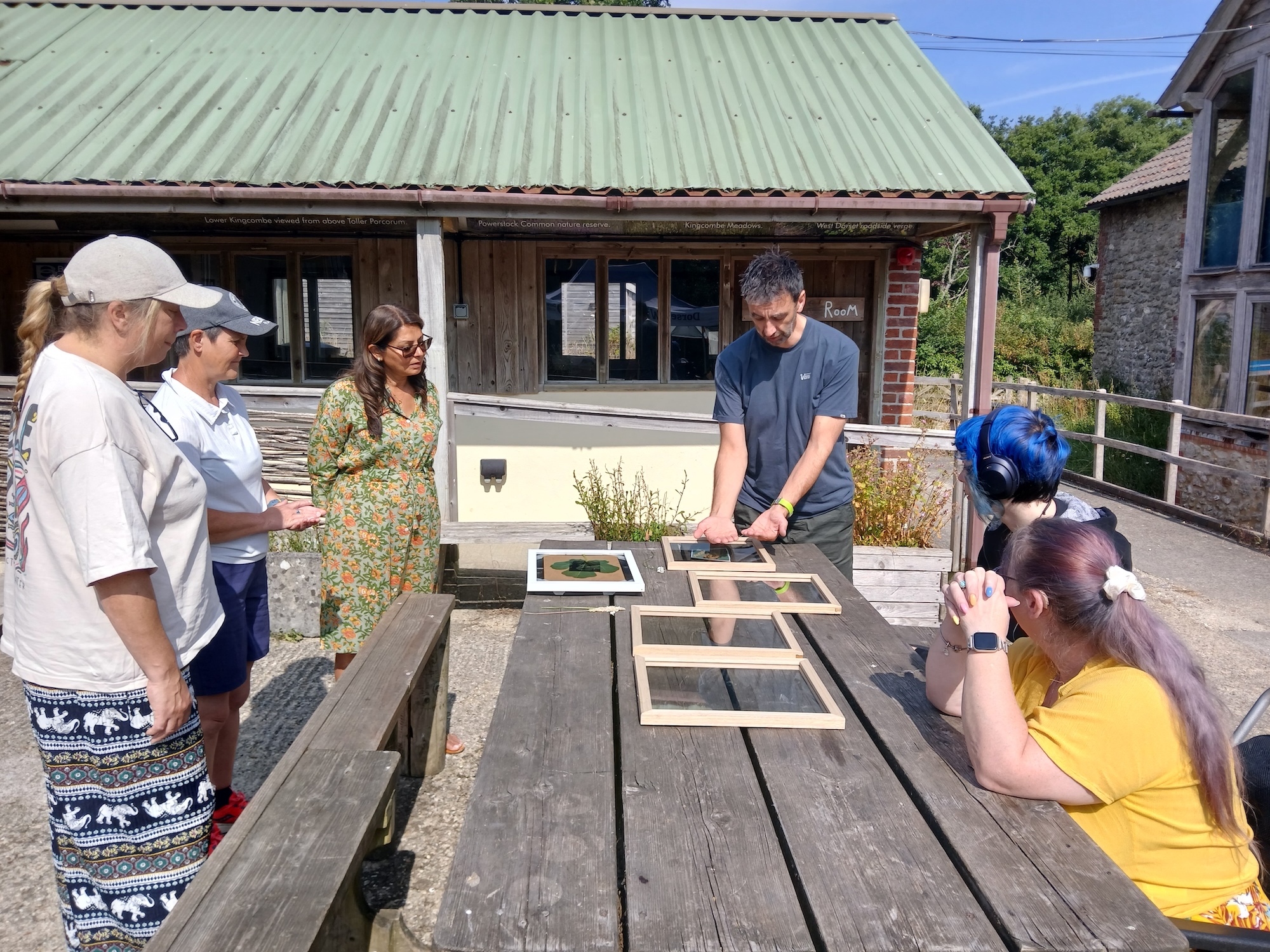 A person arranges framed items on a wooden picnic table while four people stand and sit nearby outside a building with a green roof.