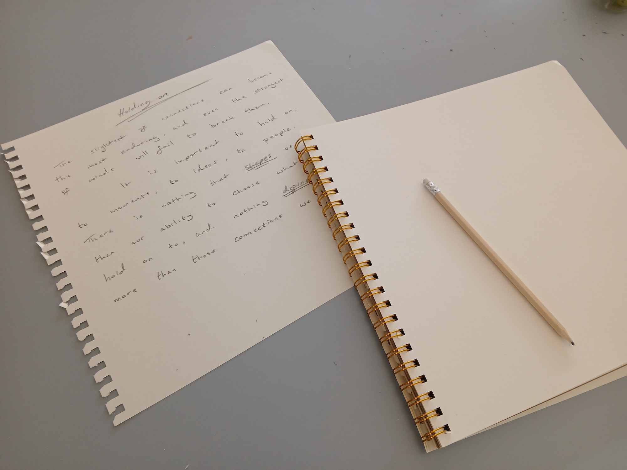 An open spiral-bound notebook with handwritten notes on the left page and a pencil resting on the blank right page, placed on a grey tabletop.