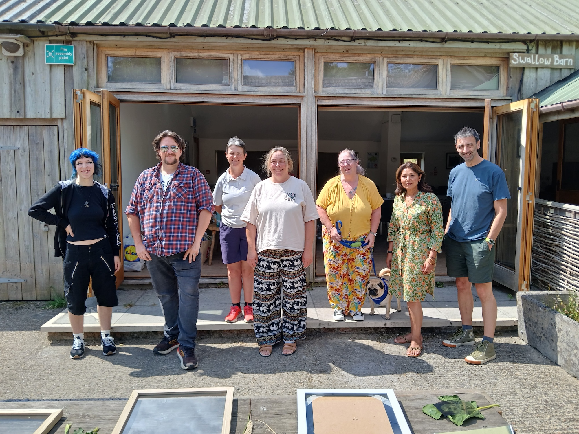 Seven people stand outside a wooden building with open doors and a corrugated roof, with potted plants and display boards in the foreground.