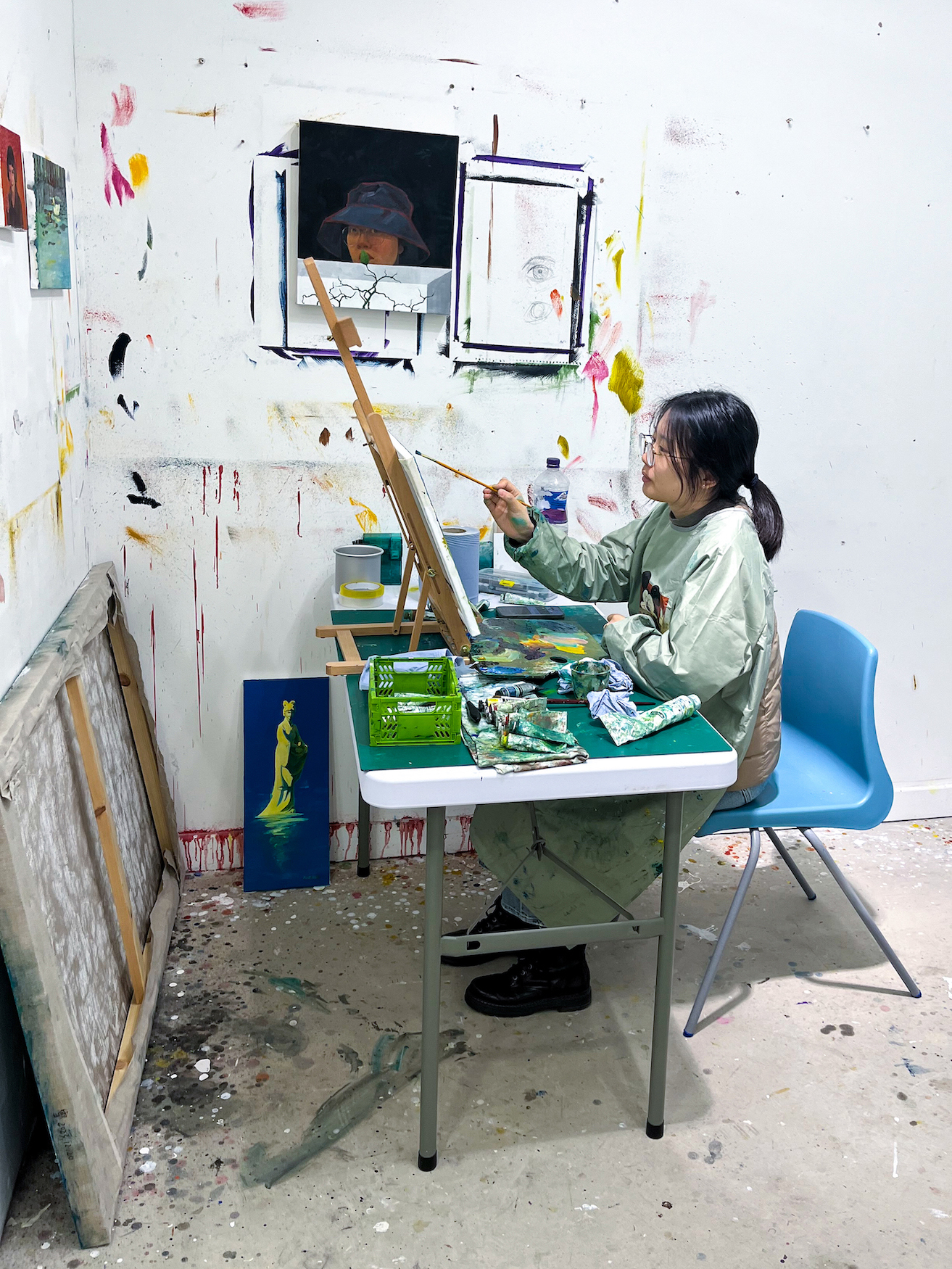 A student painting in a messy studio space with paint on the wall and floors.