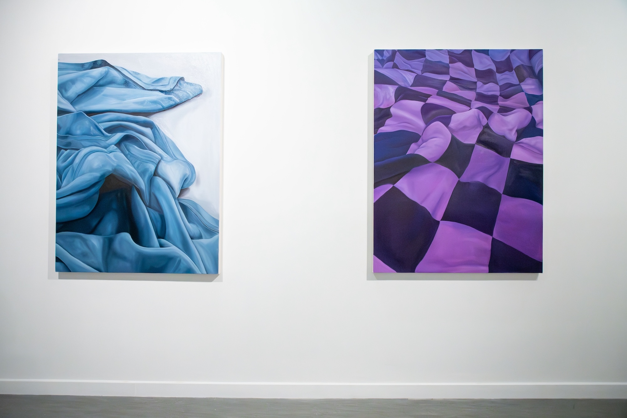 Two artworks on display in a gallery. The left painting depicts a bundle of pale blue fabric. The right painting shows a length of purple and blue check fabric.