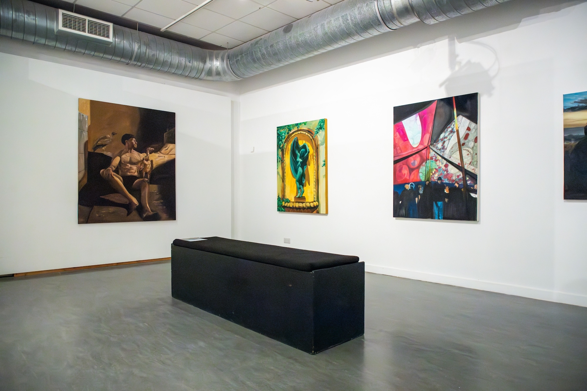 A corner of a gallery space at Lighthouse, Poole with three paintings visible hanging on the walls, a black seat in the centre.