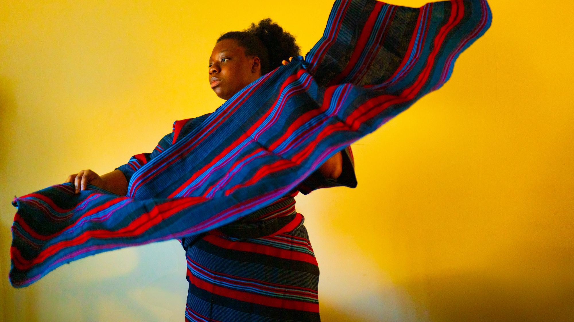 A model draped in a long blue, red and purple garment and holding a piece of fabric with the same pattern, poses for a photograph against a golden yellow background.