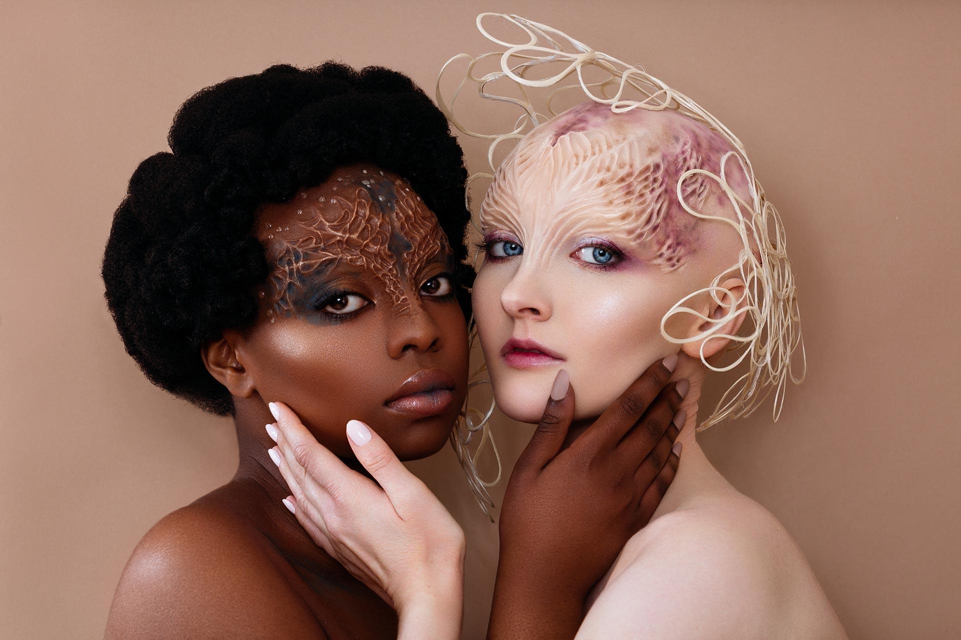 Two people with theatrical make-up and prosthetics look towards the camera touching one another's faces.