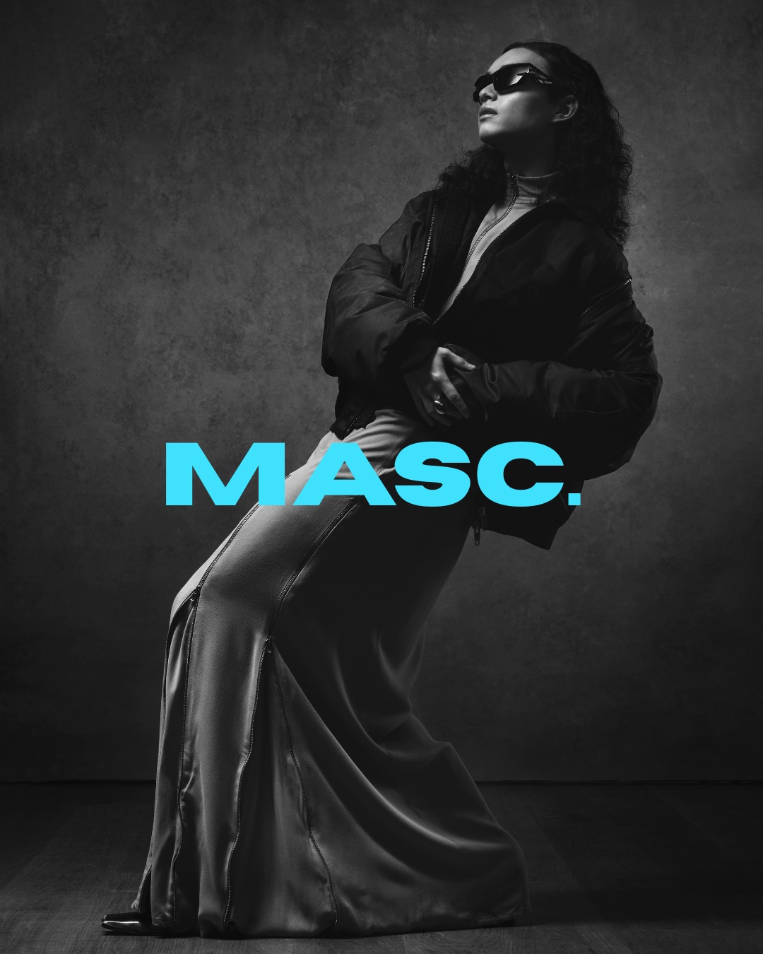 A woman in sunglasses and a long dress leans back against a dark studio backdrop, with the word “MASC.” in bright blue across the lower centre.